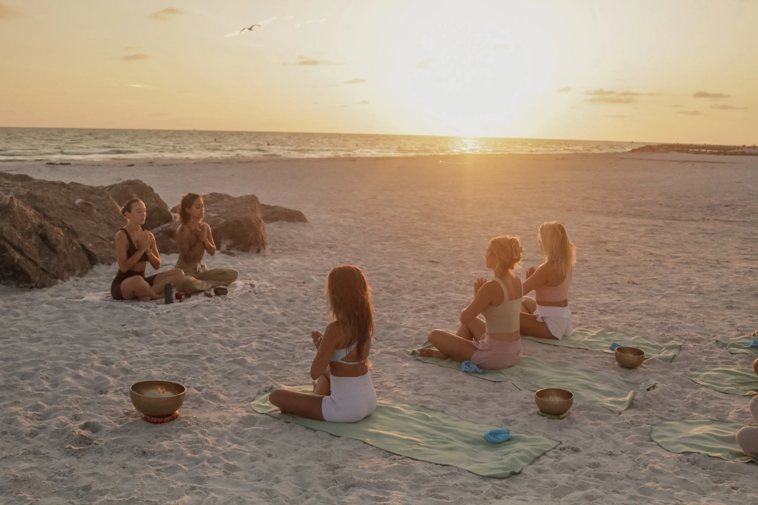 A group of people meditating on the beach during sunset, sitting on mats with bowls in front of them, near rocks with the ocean and setting sun in the background.
