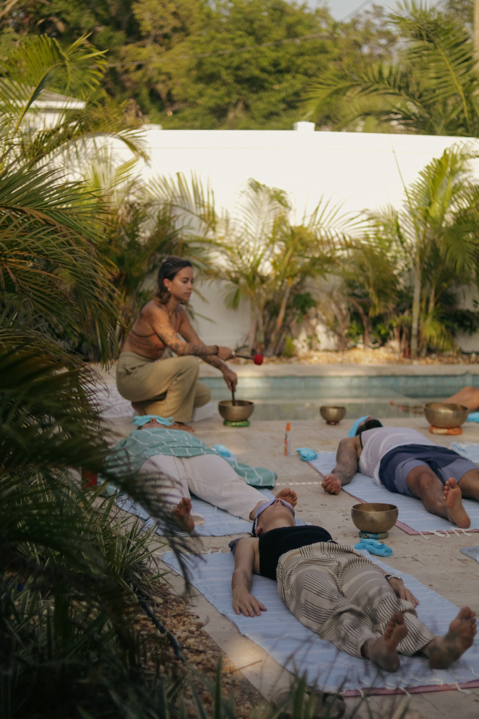 People participating in an outdoor sound healing or meditation session near a pool, surrounded by lush green plants and trees.