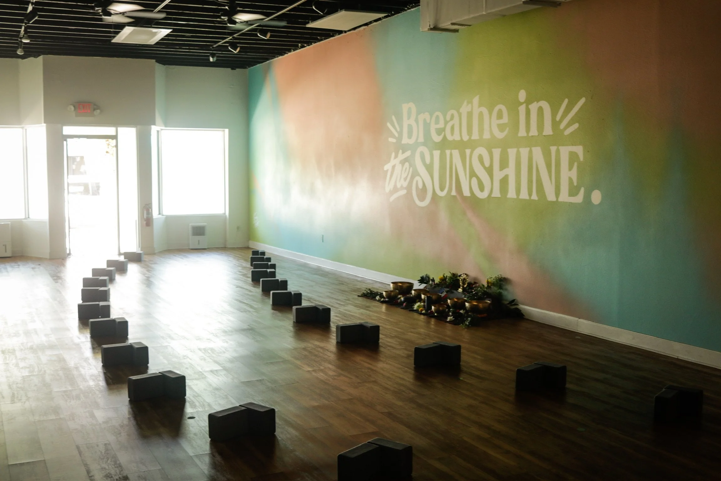 Empty yoga or meditation space with black blocks arranged in a line on wooden floor, a colorful wall with the phrase 'Breathe in the Sunshine', and plants with bowls and flowers on the floor.