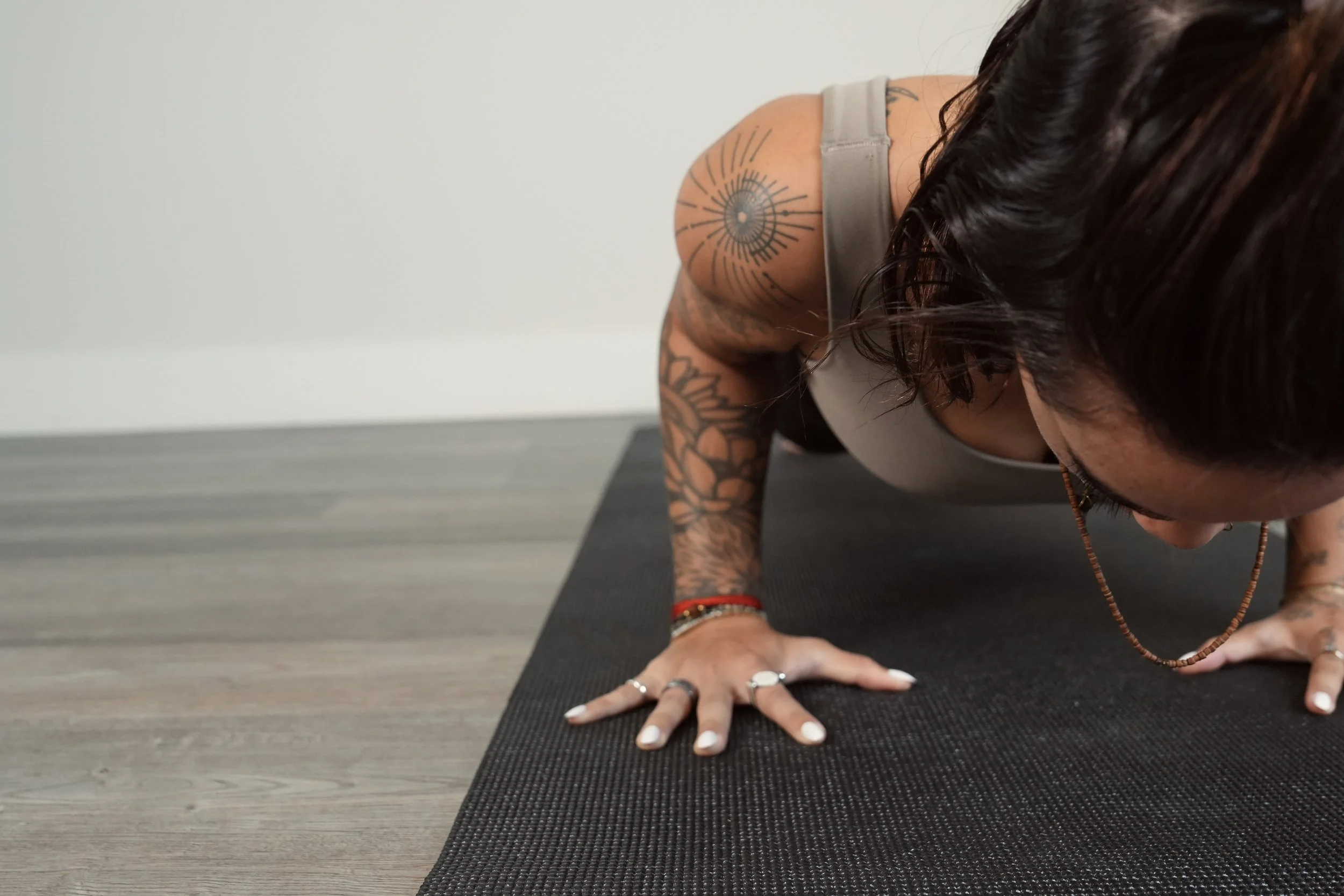 Woman with tattoos in a gray tank top doing a plank exercise on a black yoga mat on a wooden floor.
