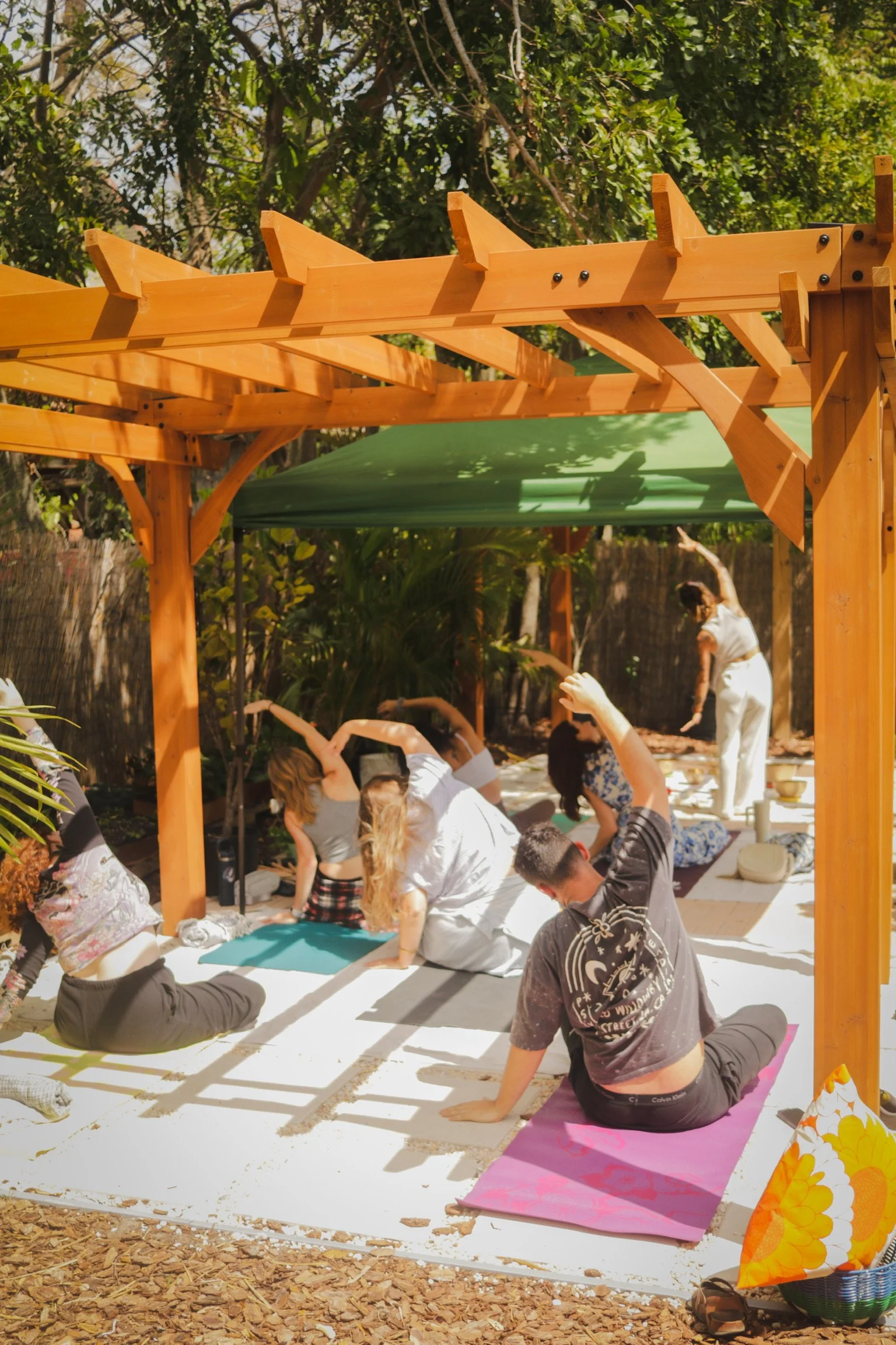 People practicing yoga outdoors under a wooden pergola on a sunny day.