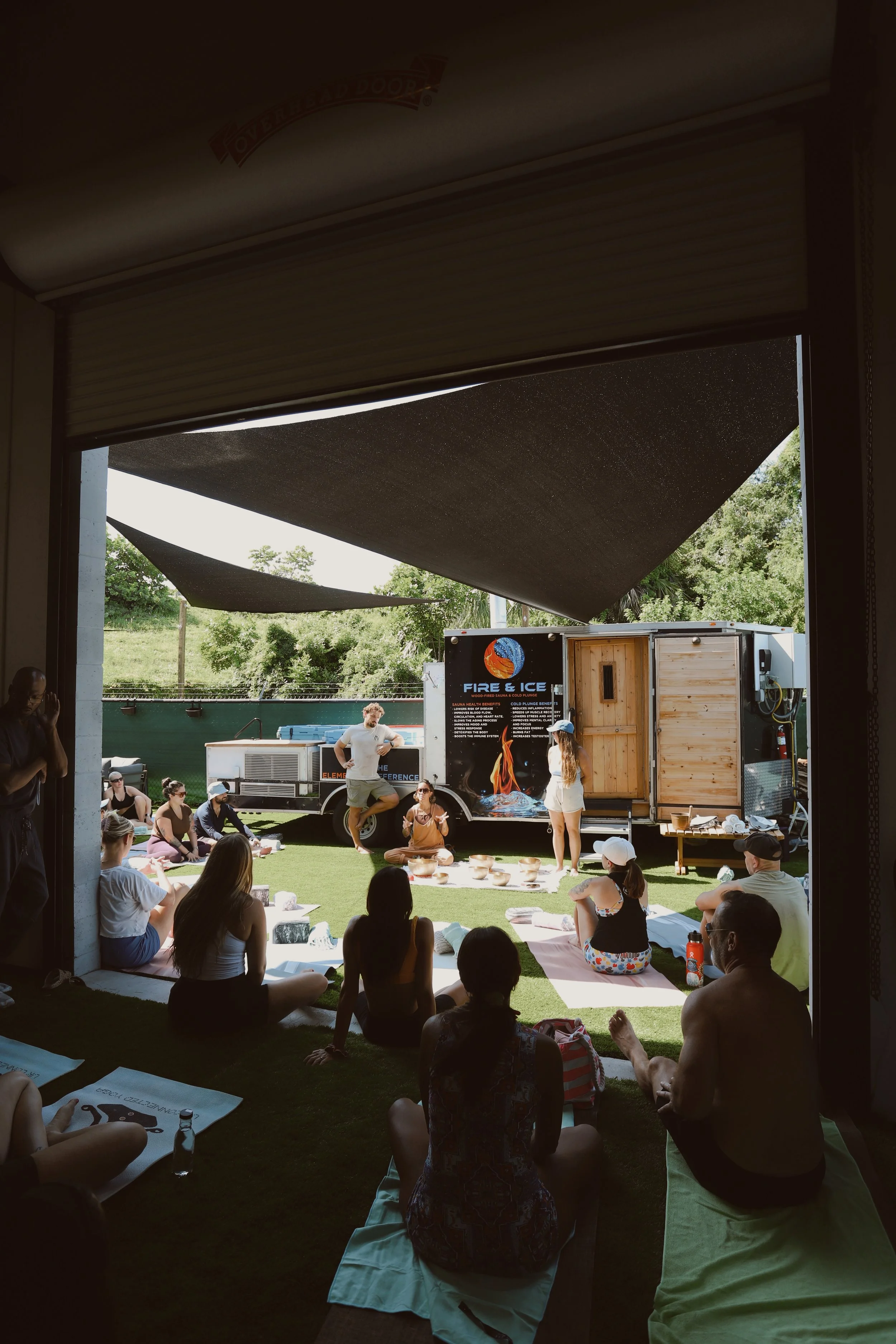People attending a yoga or meditation class outdoors next to a food truck with a "Fire & Ice" sign, sitting on mats in a shaded area and others on the grass.