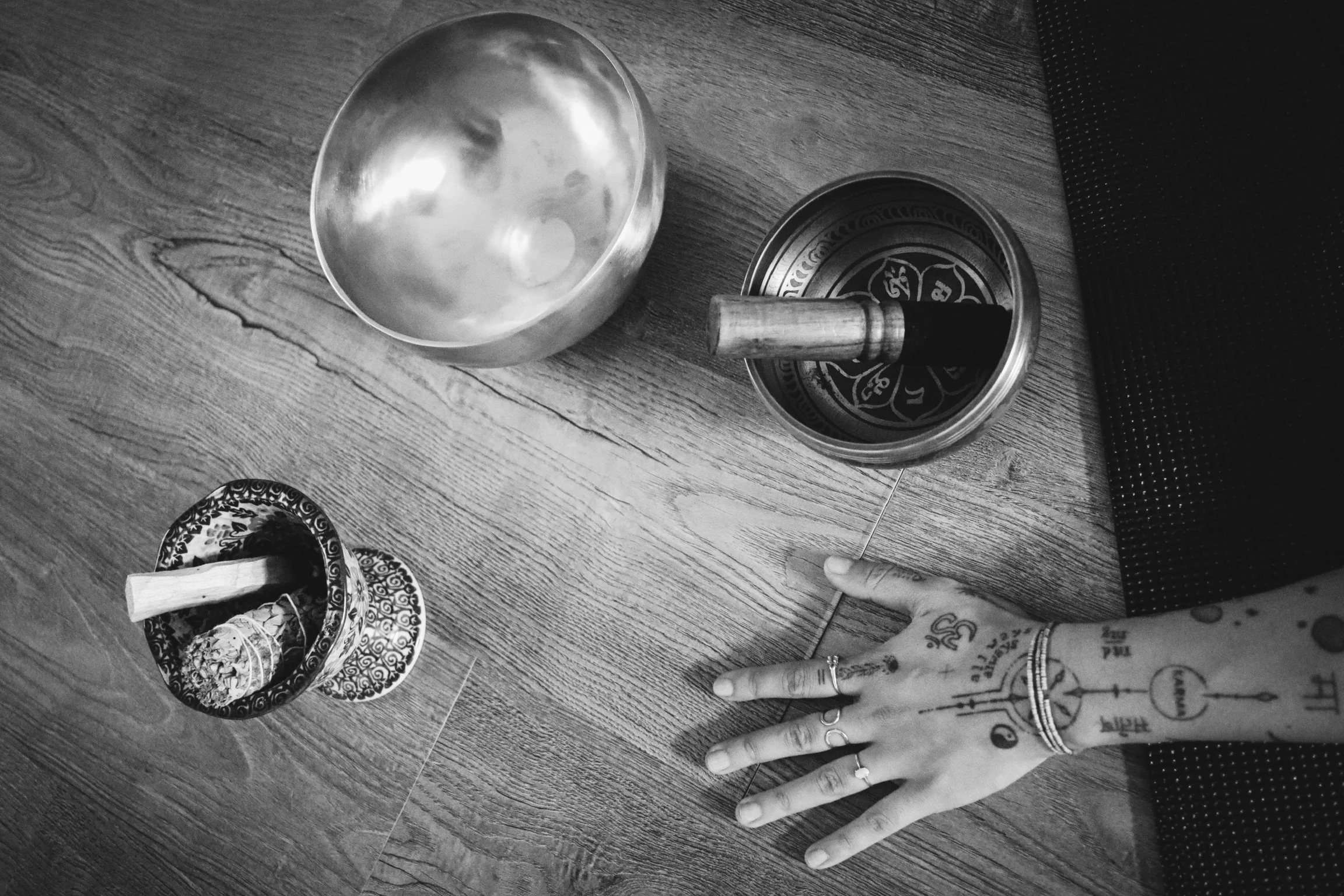 A hand with tattoos and rings resting on a wooden surface, surrounded by decorative bowls and a singing bowl with a wooden mallet.