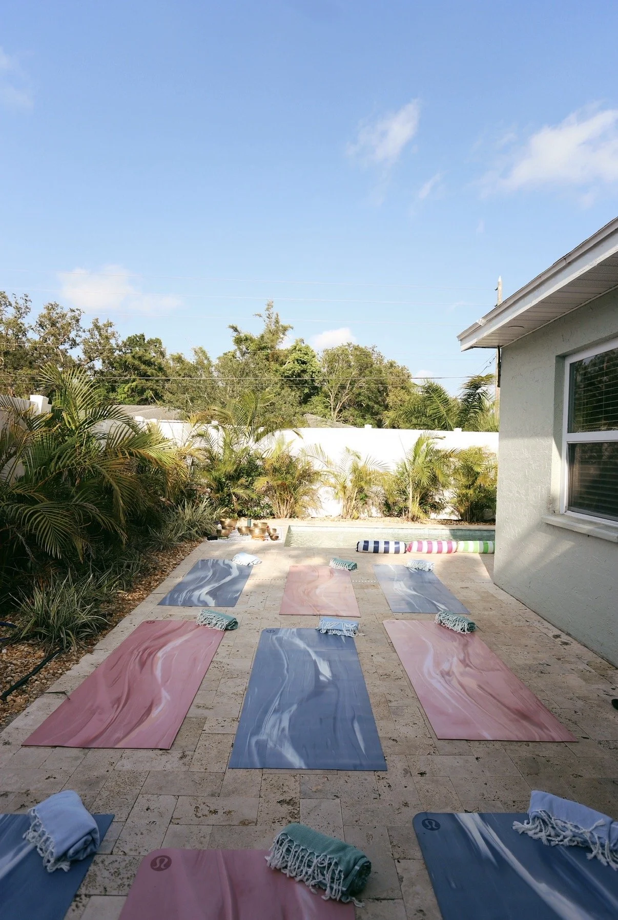 Outdoor yoga setup with pink and blue mats, rolled-up towels, and a colorful striped inflatable pool float, on a paved patio next to a house with a window and a garden with palm plants, under a clear blue sky.