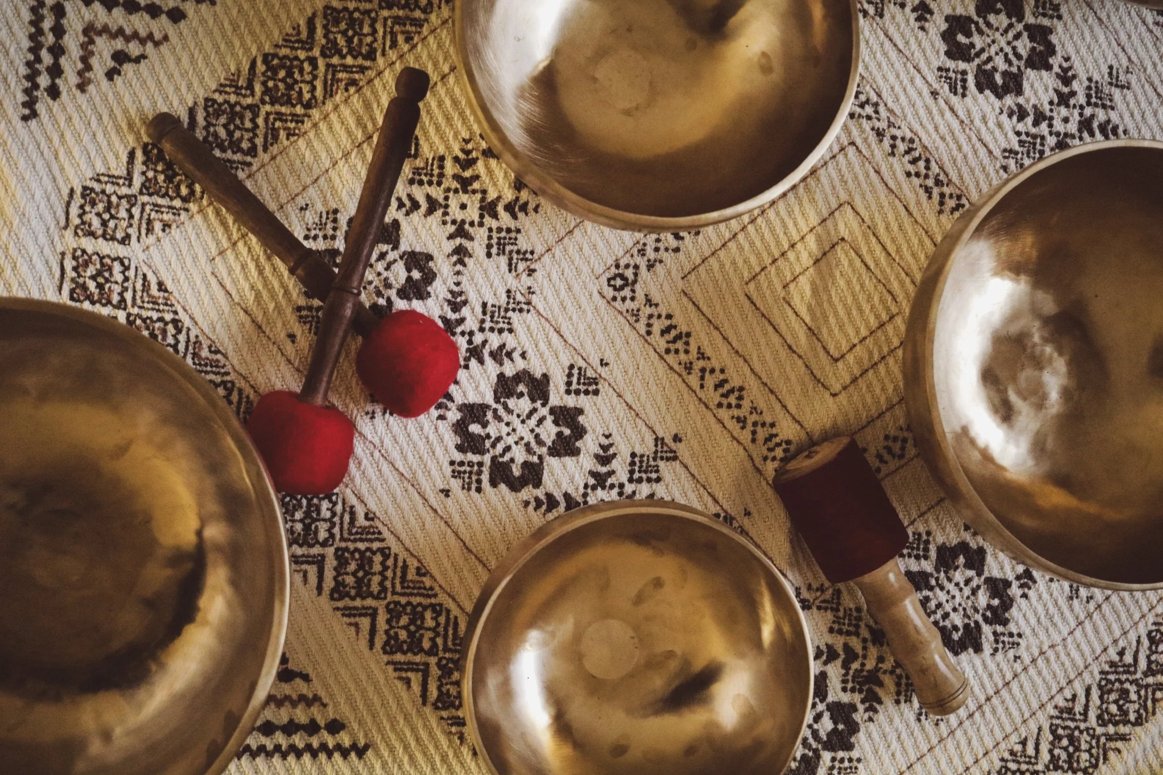Four metallic hand cymbals, a wooden mallet with two red felt balls, and a small wooden stick on a patterned fabric surface.