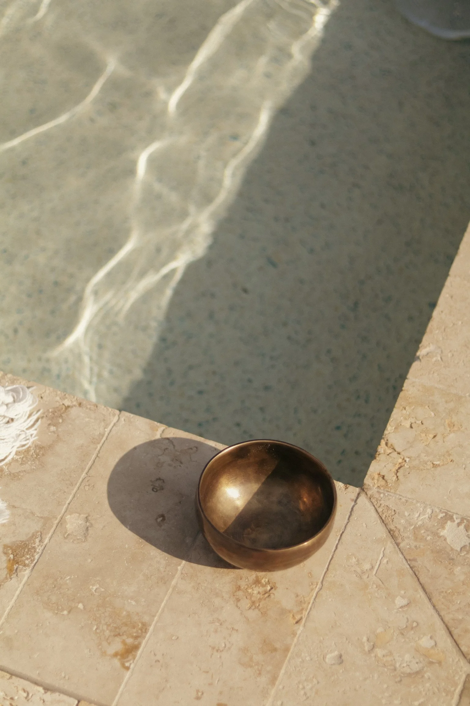 A small metallic bowl placed on a beige tiled surface outside, with shadows and sunlight, near a swimming pool with clear water.