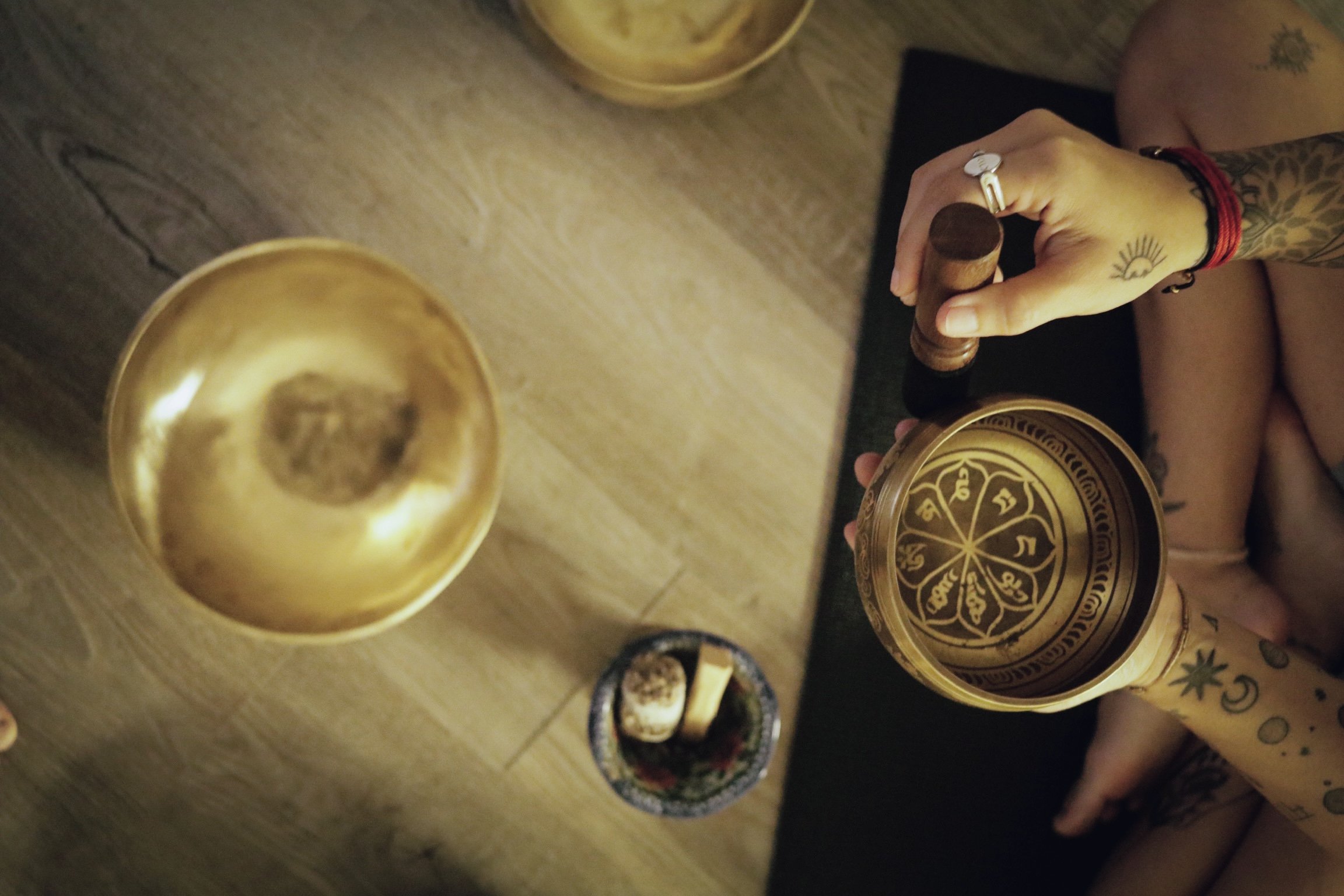 A person with tattoos on both arms holding a brass singing bowl and a wooden mallet, sitting at a wooden table with another brass singing bowl and a small decorative container on it. The setting appears to be a cozy indoor space.
