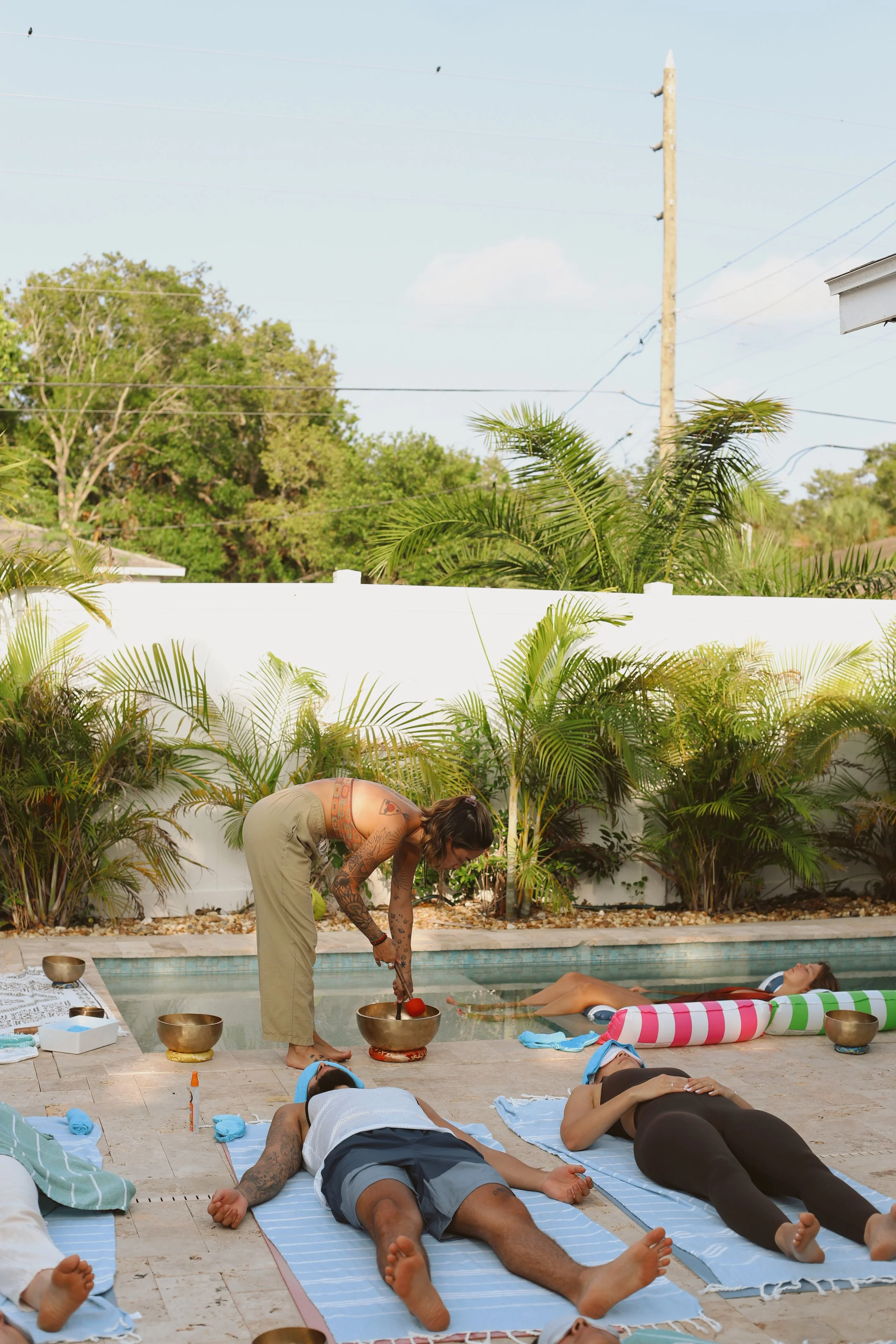 People participating in a sound bath meditation outside near a pool with tropical plants, with some lying on towels and one person, possibly the instructor, holding a singing bowl.