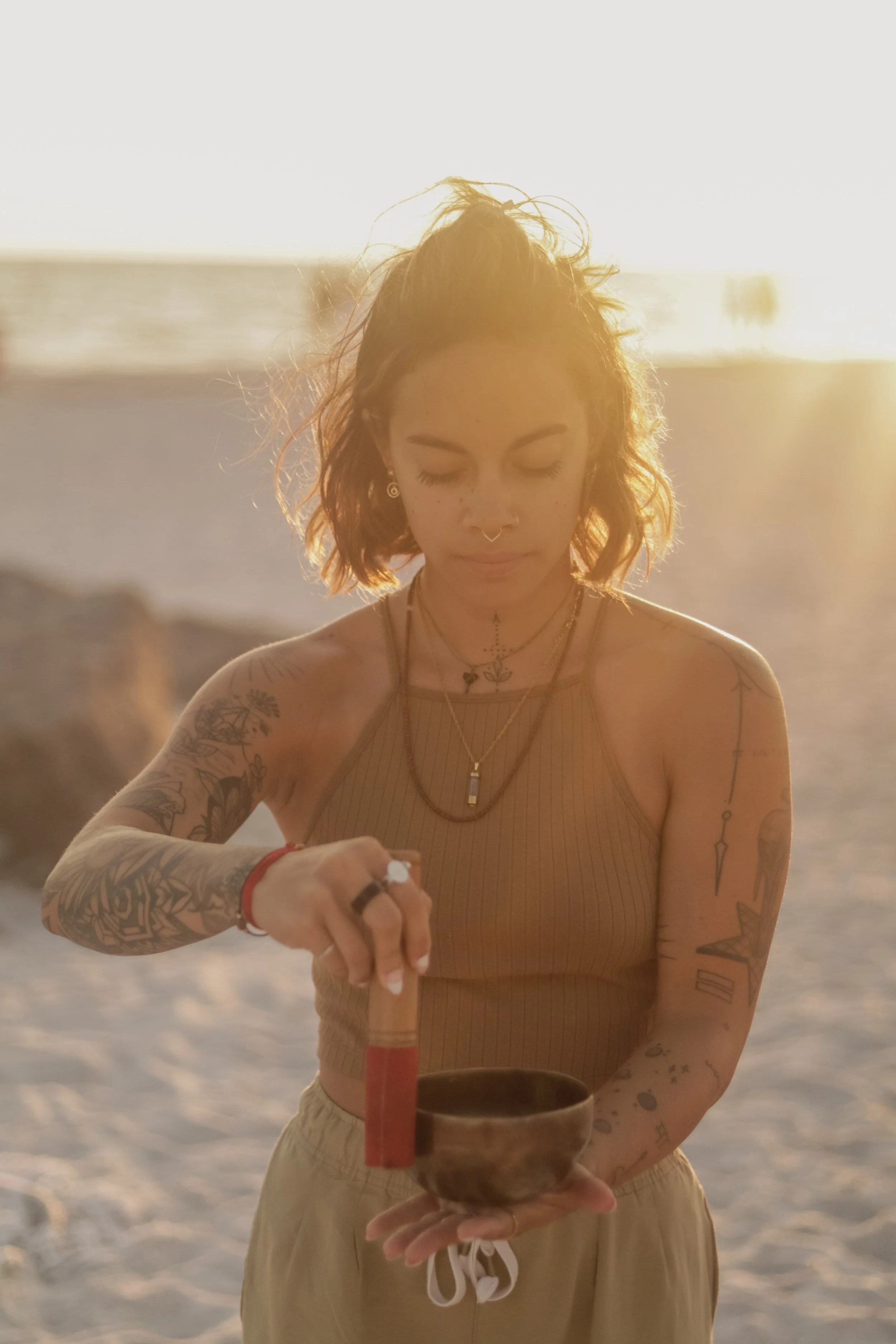 Woman with tattoos on arms holding a singing bowl and mallet on a beach during sunset