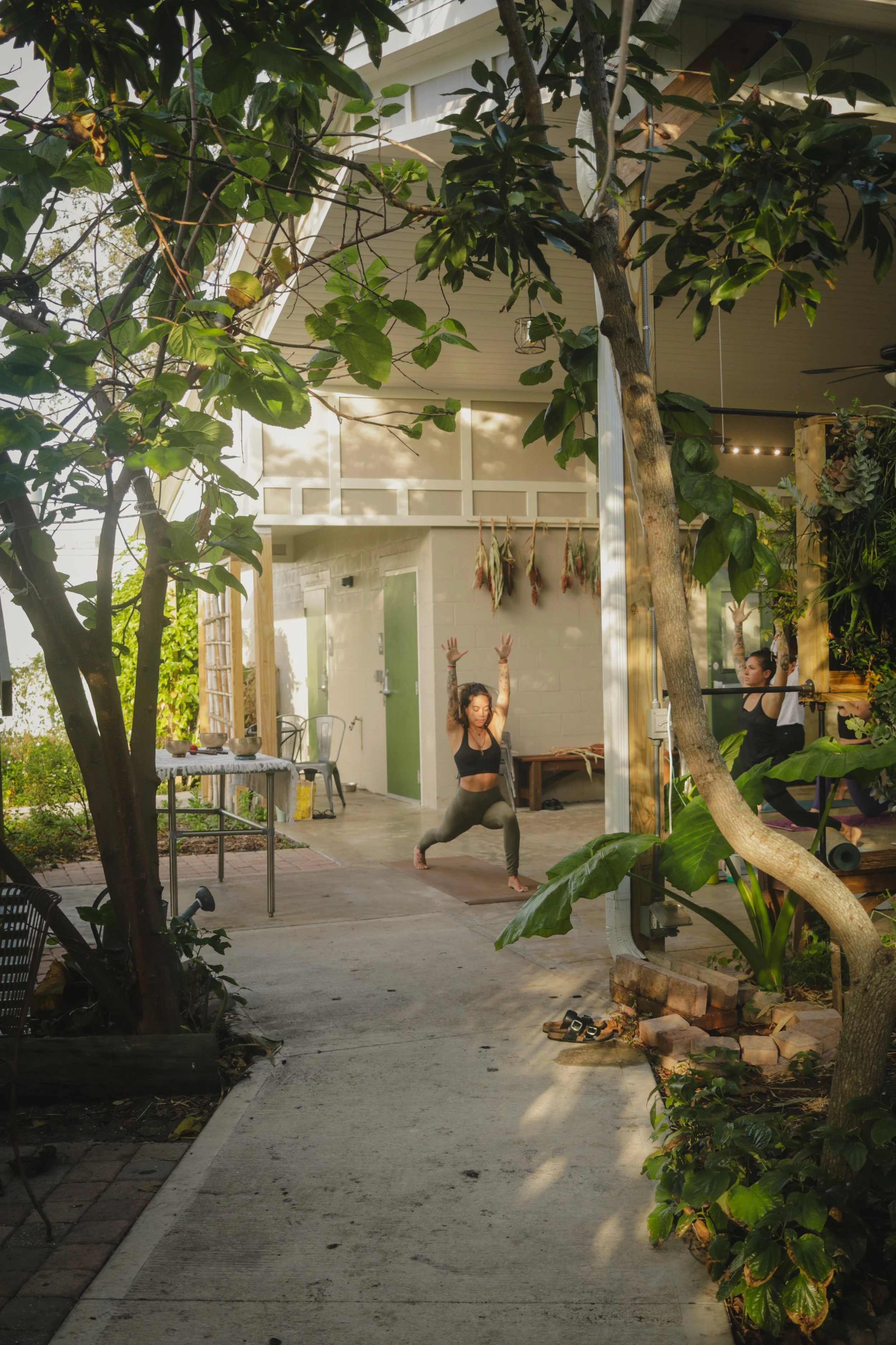 Two women practicing yoga outdoors in a garden area, one in a lunge position with arms raised, the other on a yoga mat with arms raised, surrounded by greenery and trees.