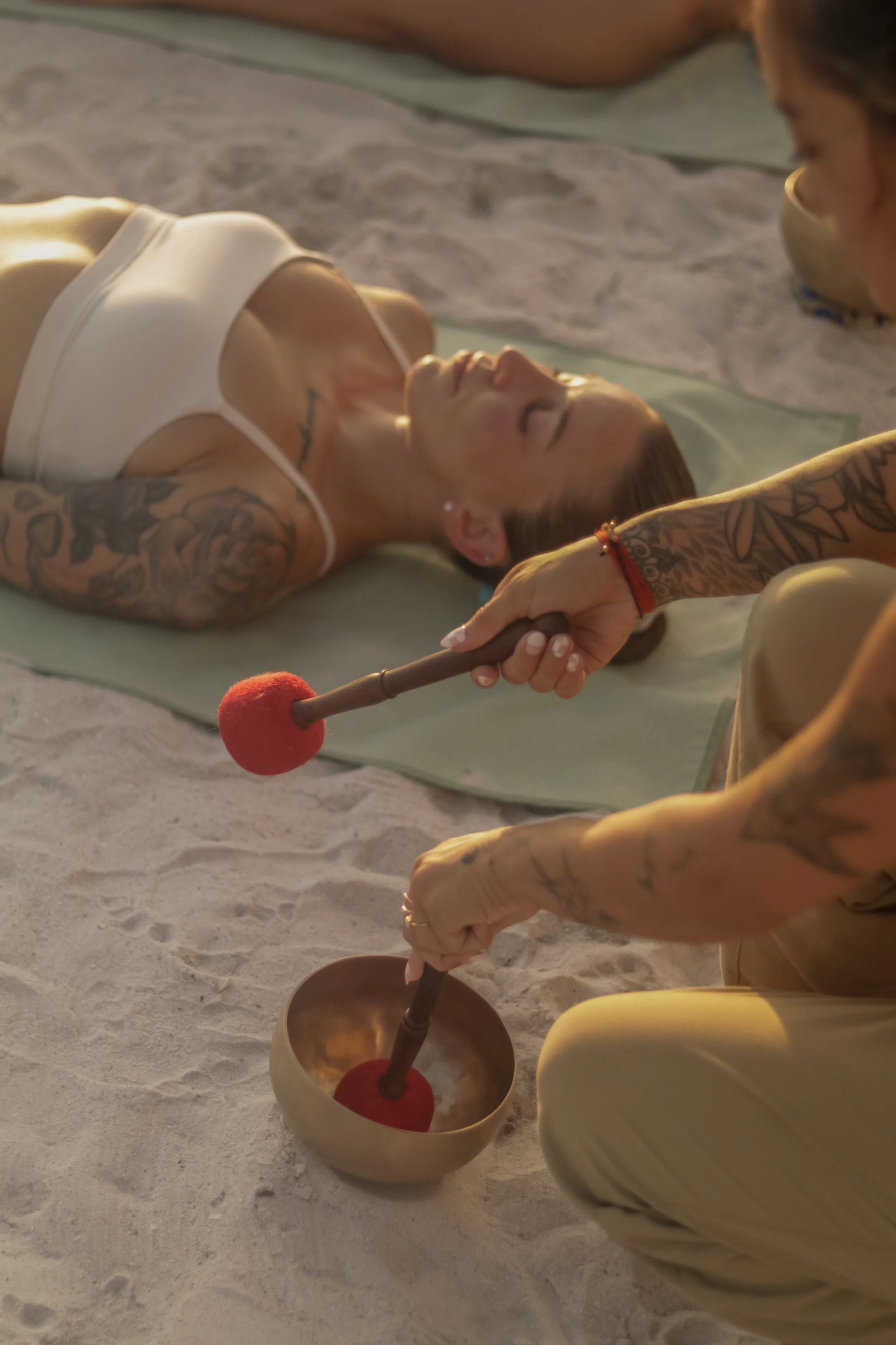 A woman with tattoos lying on a mat on the sand with eyes closed, being held by two people with matching tattoos, playing singing bowls with mallets.
