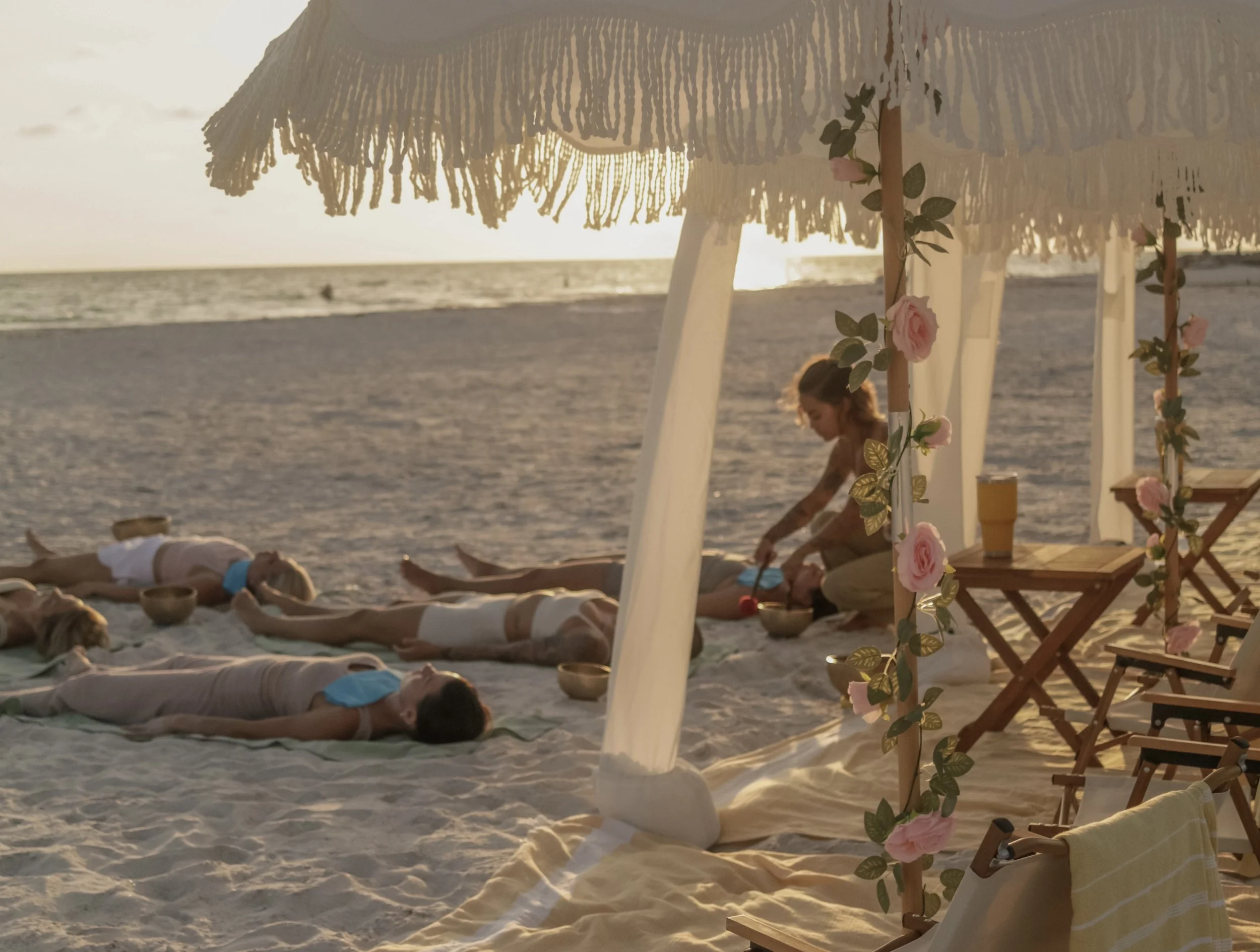 A woman practicing yoga on the beach under a decorated canopy, with several children lying on the sand nearby during sunset.