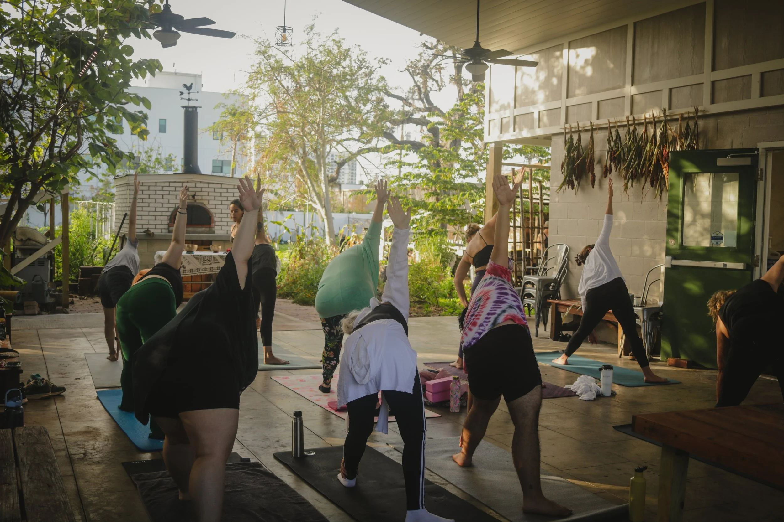 Group of people practicing yoga outdoors on mats in a shaded patio, with some participants raising one arm up and bending sideways, during daylight.