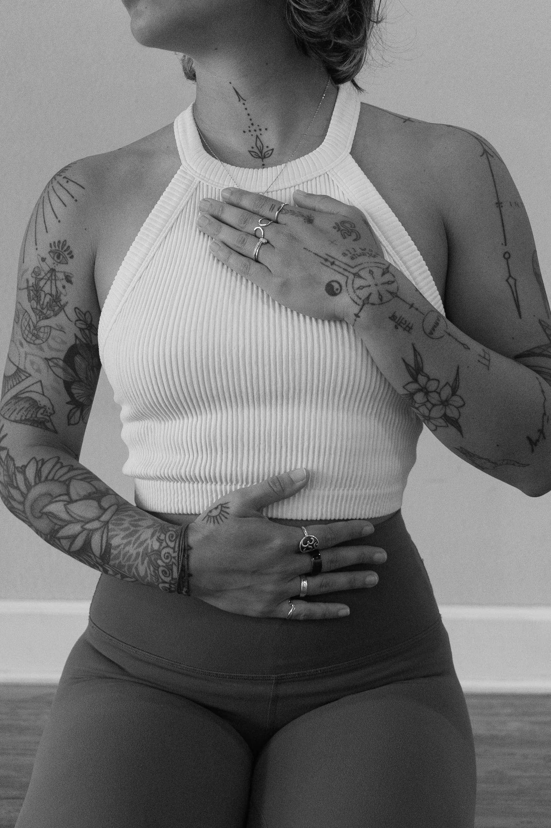 A person with tattoos on their arms, chest, and neck taking a black and white mirror selfie. They are wearing a white ribbed tank top and dark leggings, with rings on their fingers and a delicate chain necklace.