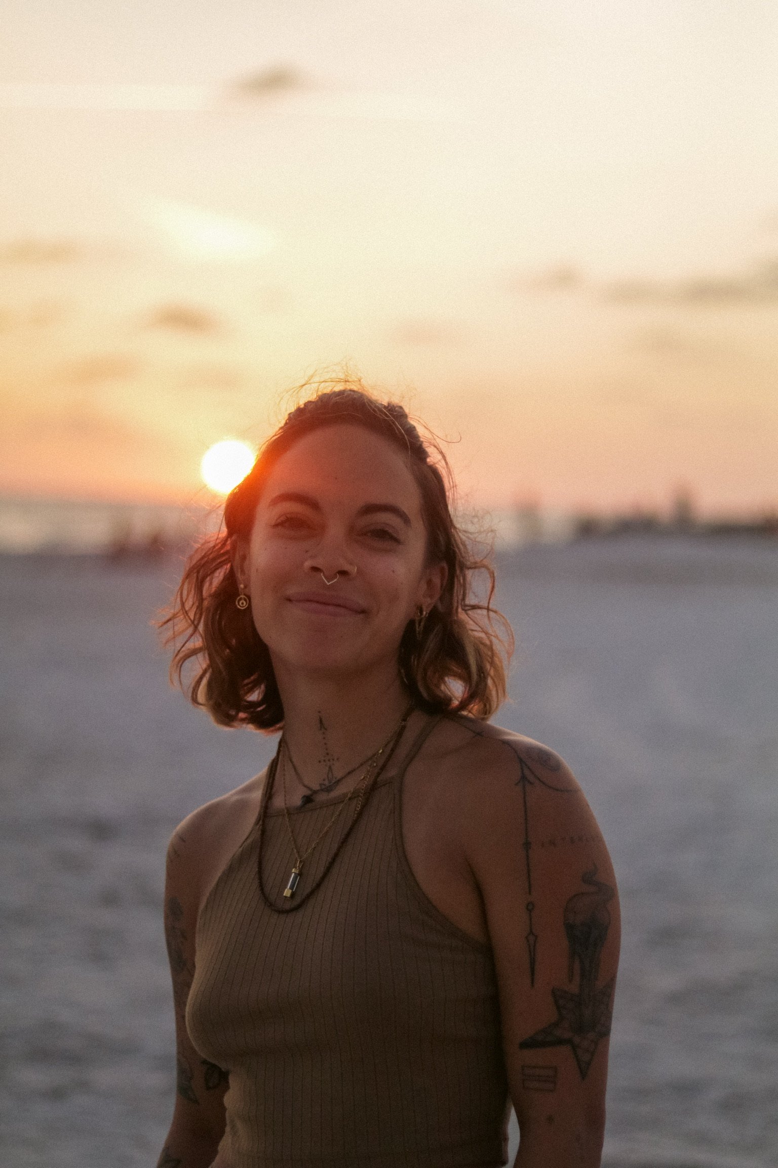 A young woman with tattoos and jewelry smiling at the camera during sunset on a beach.