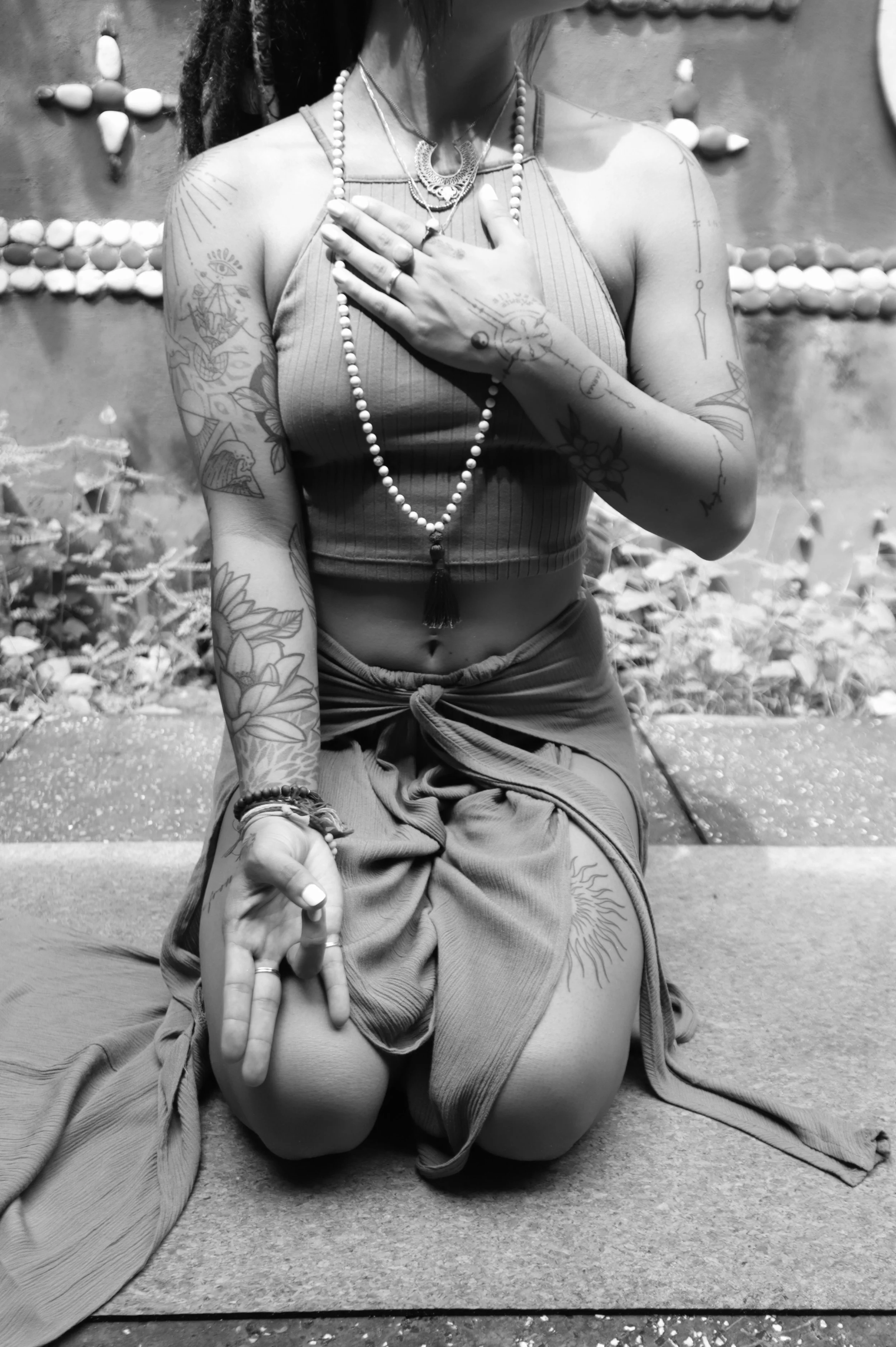 Person kneeling outdoors, wearing necklaces, with tattoos on arms and abdomen, one hand on chest and the other resting on knee, in front of a decorative wall with shell designs, black and white