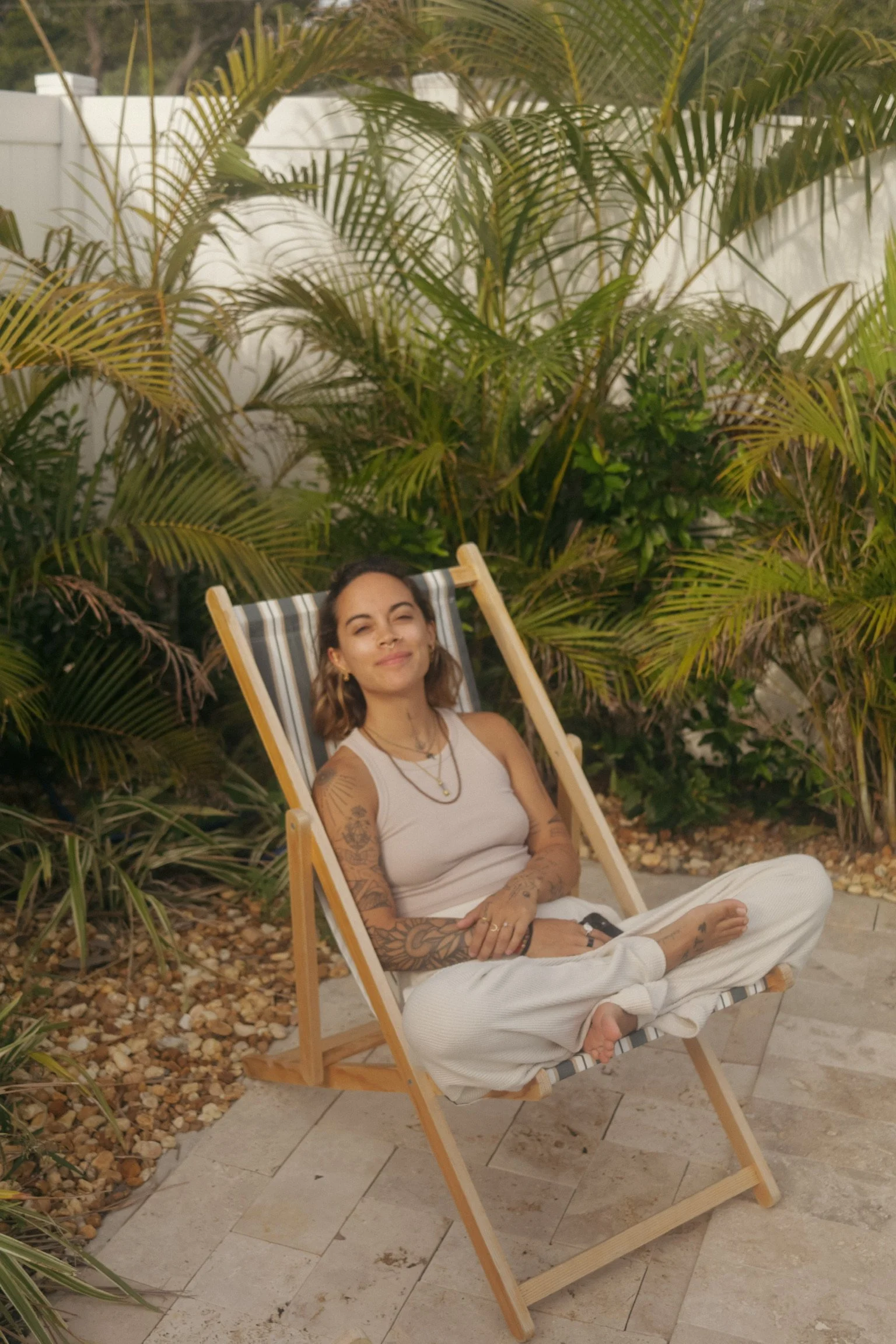 A young woman with tattoos relaxing in a striped lounge chair outdoors in a lush, tropical setting.