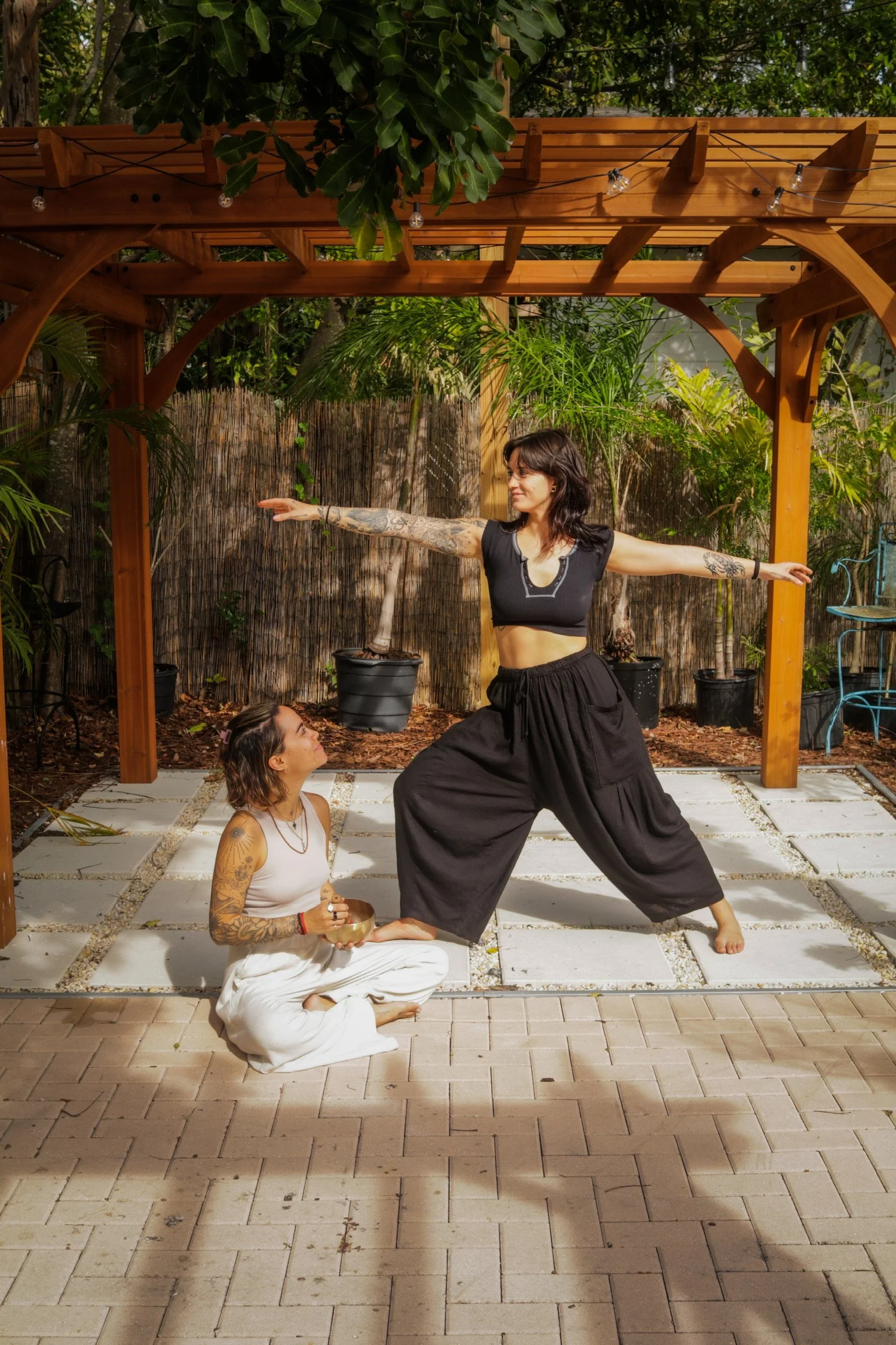 Two women practicing yoga outdoors under a wooden pergola, with one woman kneeling and holding a singing bowl, and the other woman standing with arms outstretched in a yoga pose, surrounded by greenery and potted plants.