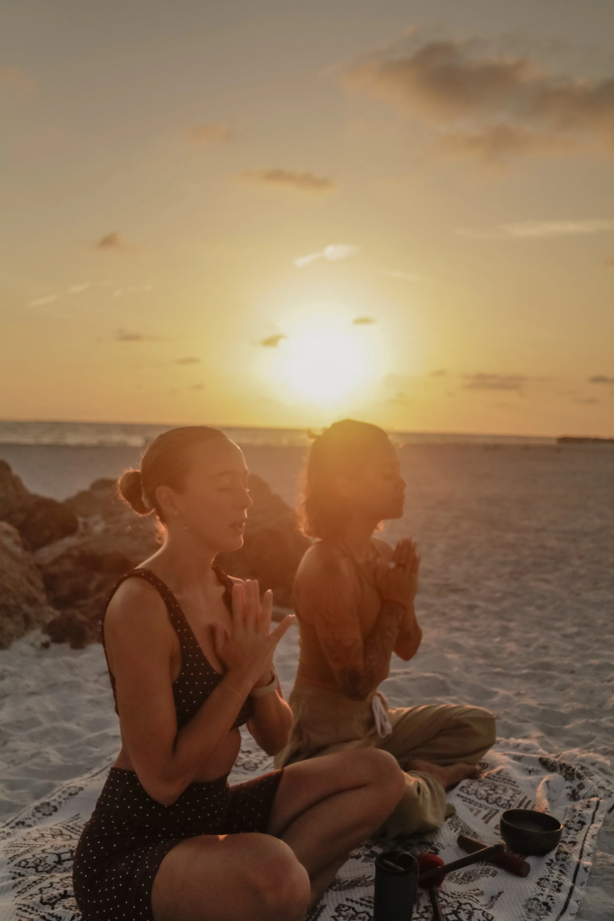 Two women practicing meditation or yoga at sunset on a beach, sitting cross-legged on a blanket with their eyes closed and hands in prayer position.