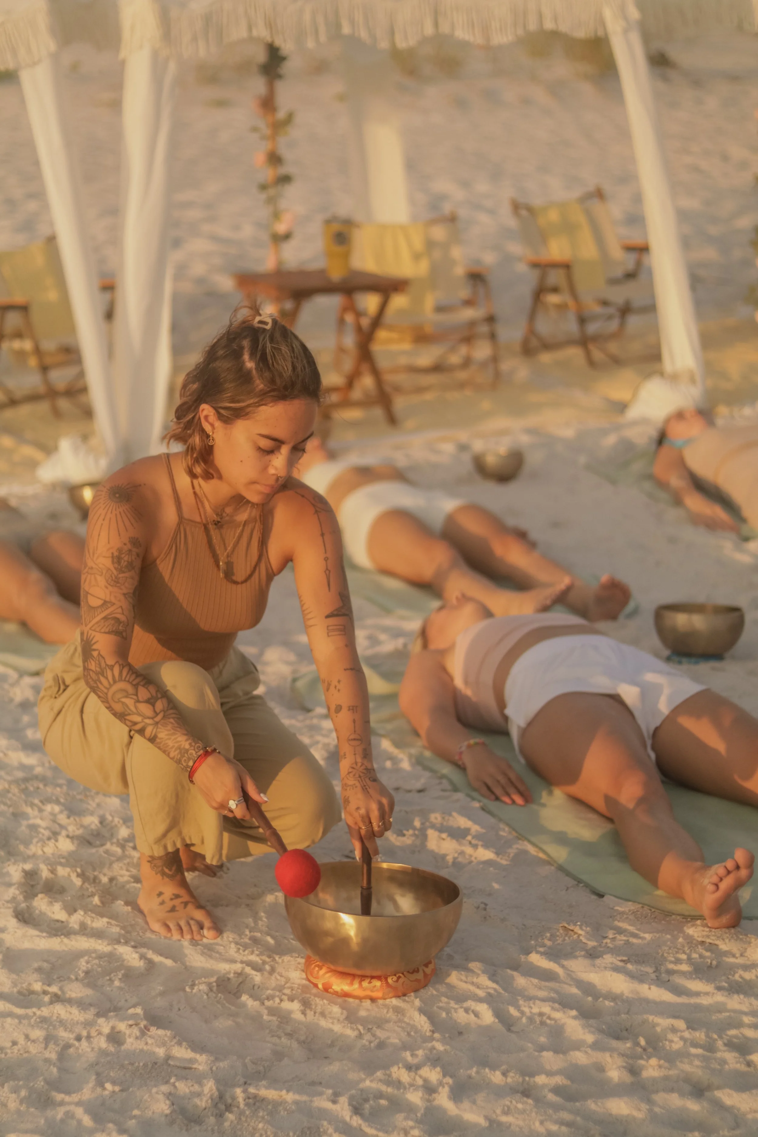 A woman with tattoos on her arms kneels on the sand, playing a singing bowl with a mallet during a beach meditation session at sunset. Several people are lying on yoga mats in the background, and chairs and tables are set up under a canopy.