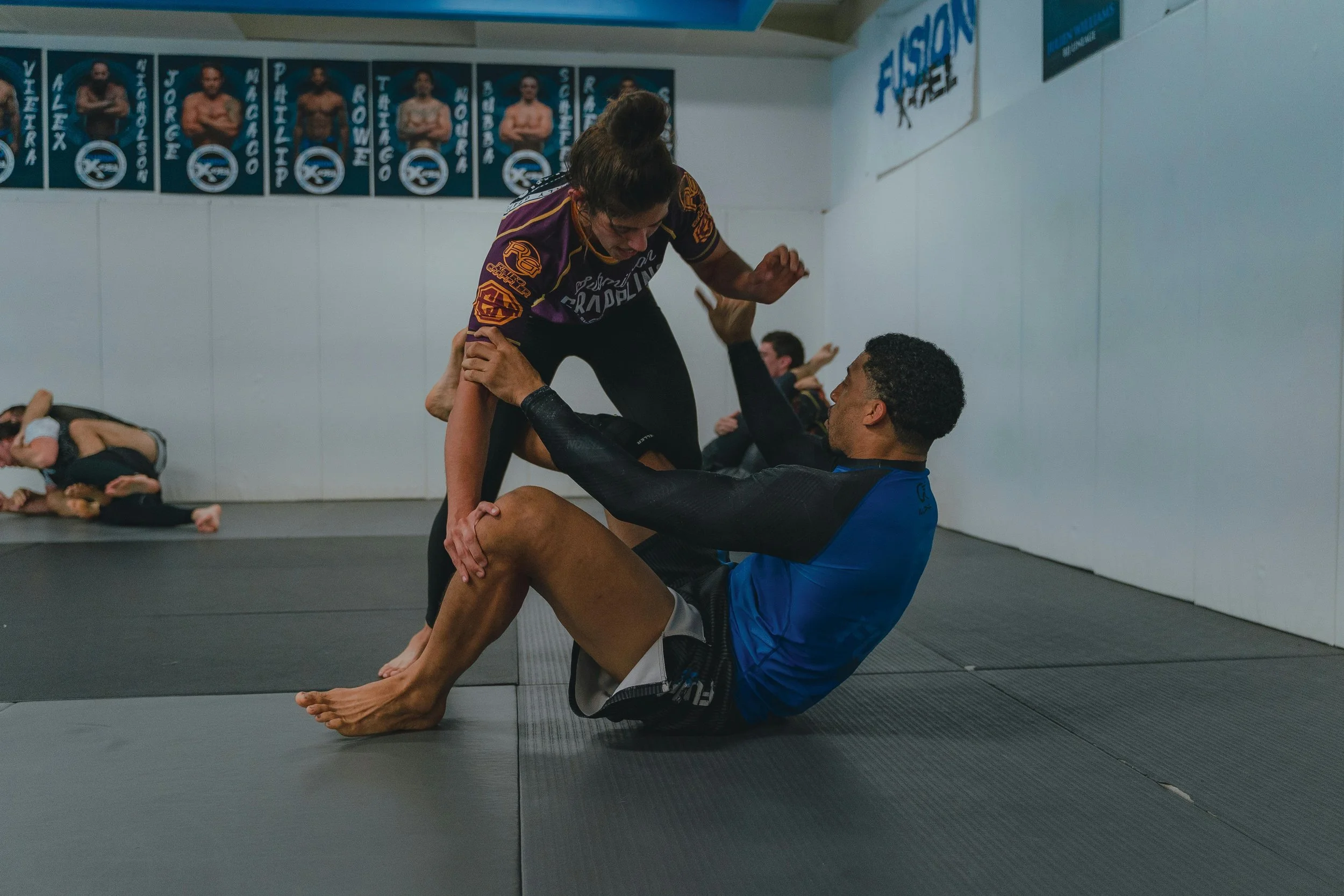 Two Brazilians practicing Brazilian Jiu-Jitsu on a black mat, with one woman in a purple gi and a man in a blue gi engaging in a grappling move, inside a martial arts gym.