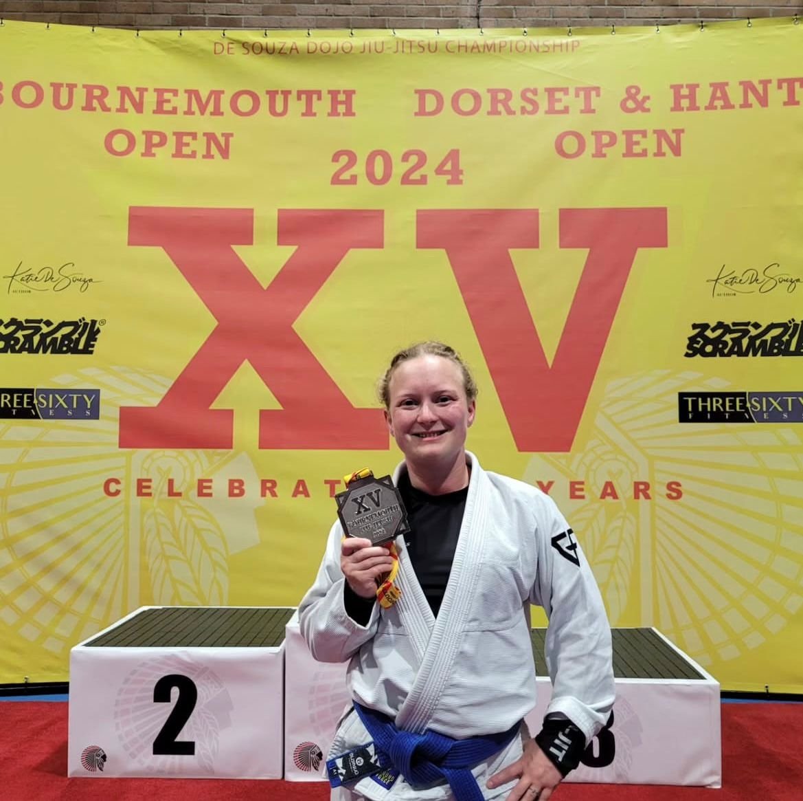 Young woman in a Brazilian Jiu-Jitsu gi holding a silver medal stands on a podium with a large yellow banner in the background. The banner promotes the 15th De Souza Dojo Jiu-Jitsu Championship, celebrating 25 years, scheduled for 2024, with references to Bournemouth, Dorset, and Hants.