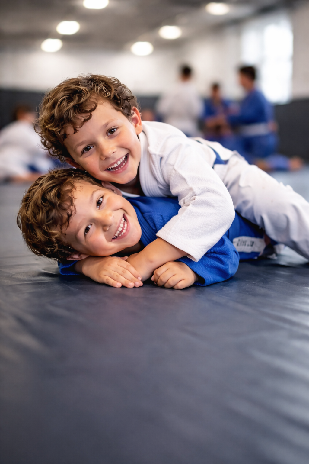 Two young boys with curly hair smiling and playing together on a martial arts training mat, wearing gi uniforms, with other students in gi in the background.