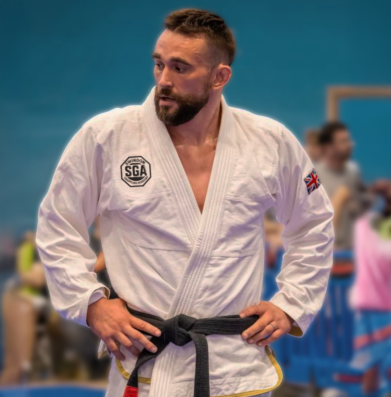 A man wearing a white judo gi with patches, including a Swindon SGA patch and a UK flag patch, standing with his hands on his hips at a martial arts training facility.