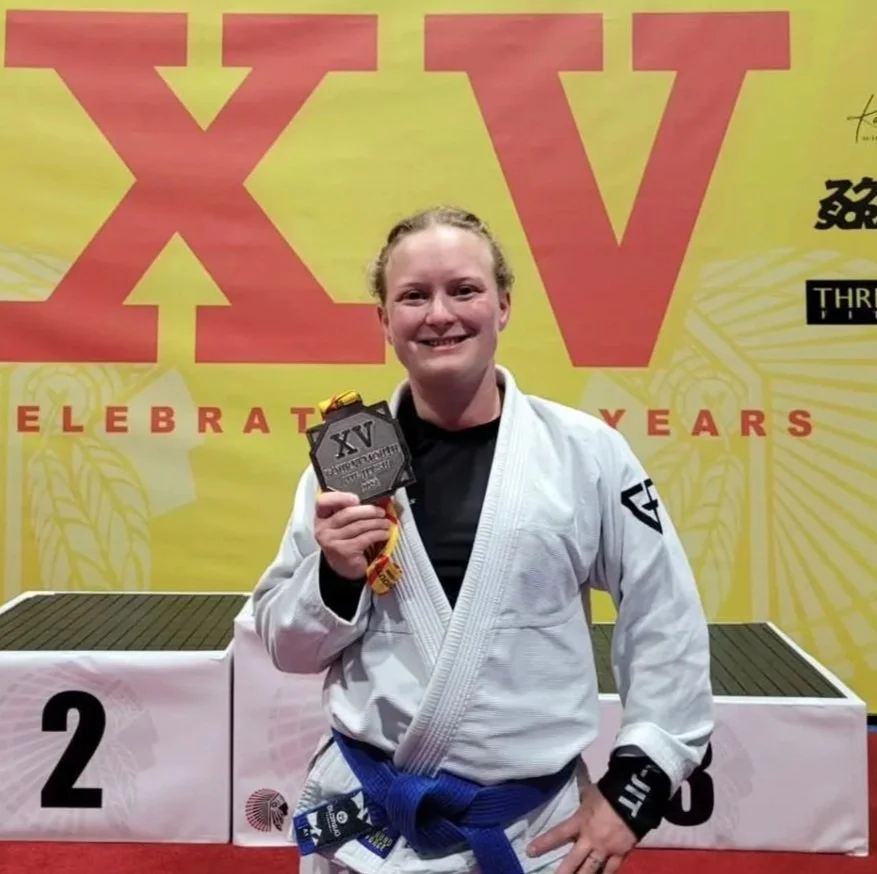 Young woman holding a medal, standing on a podium with a 2nd place position, in front of a large yellow and red banner celebrating 15 years, wearing a white martial arts gi and a blue belt.