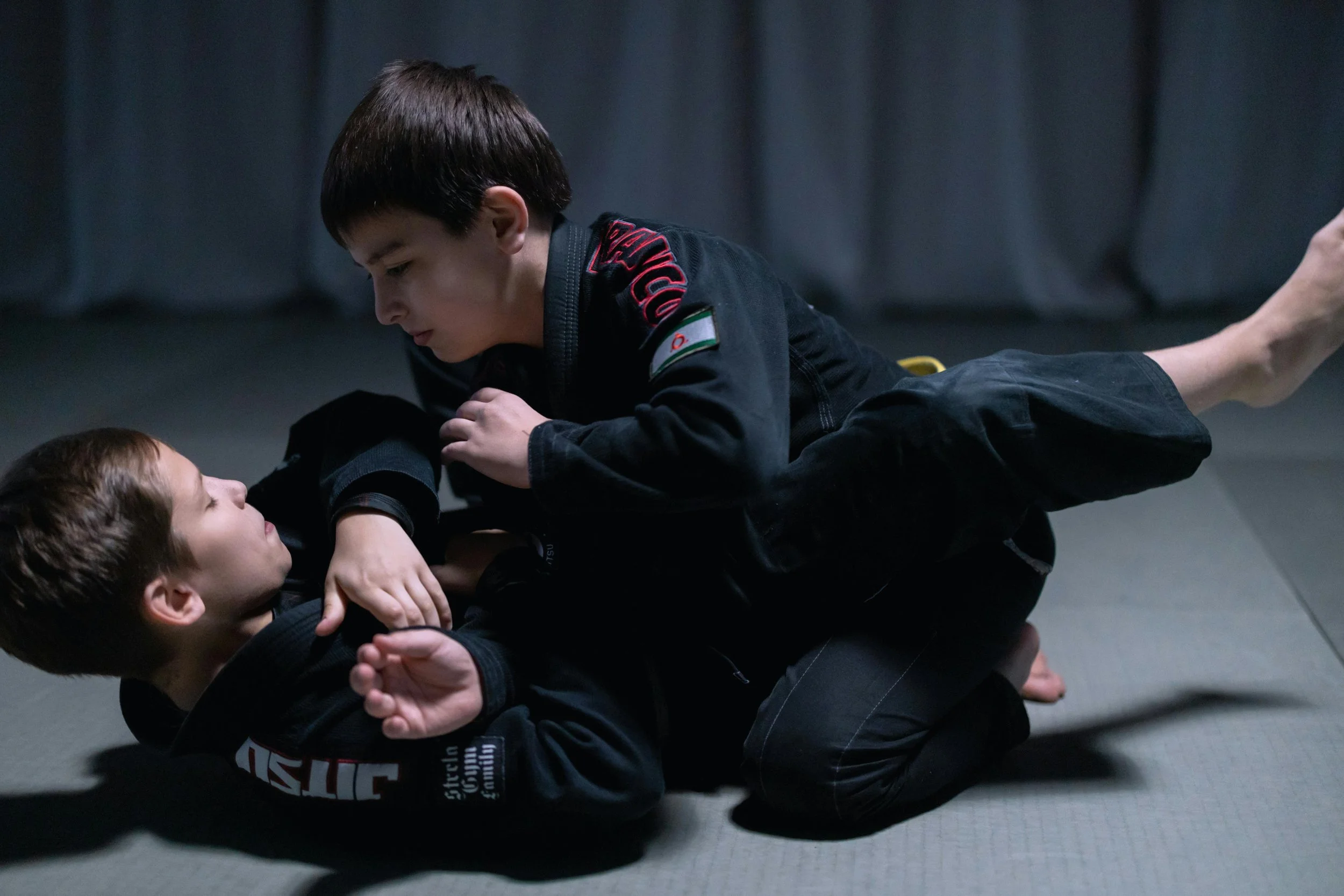 Two children practicing Brazilian Jiu-Jitsu on a mat, with one child on top and the other underneath, both wearing martial arts gi uniforms.