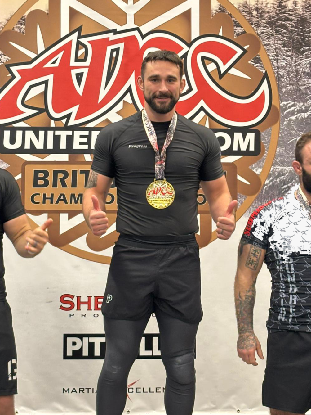 A man with a beard and short hair standing on a podium, wearing a black athletic shirt and leggings, with a medal around his neck, giving two thumbs up. Behind him is a logo for the UK Advanced Martial Arts Championships, with a snowy forest in the background.