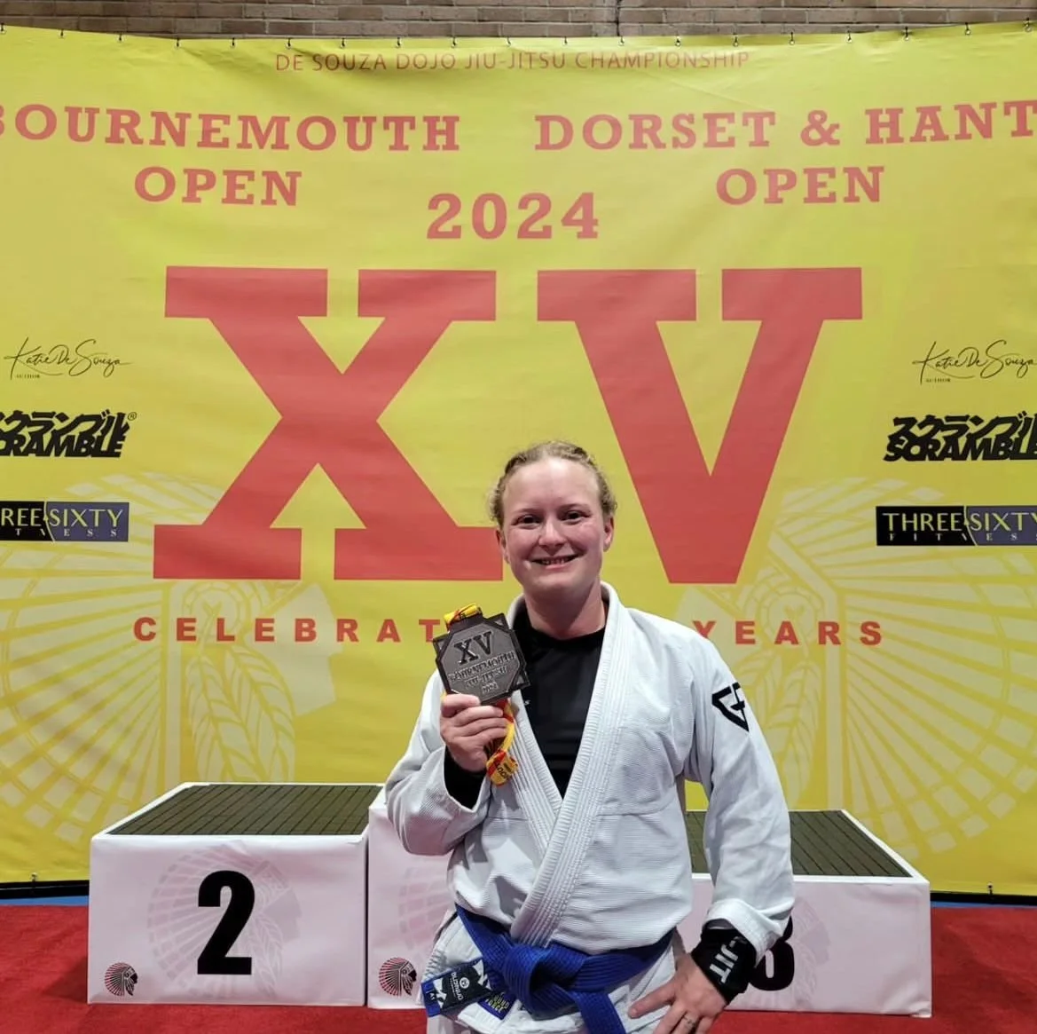 A woman in a white Jiu-Jitsu gi holding a medal, standing on a podium at the 15th Bournemouth Dorset & Hampshire Jiu-Jitsu Championship in 2024, with a yellow banner background displaying event information.