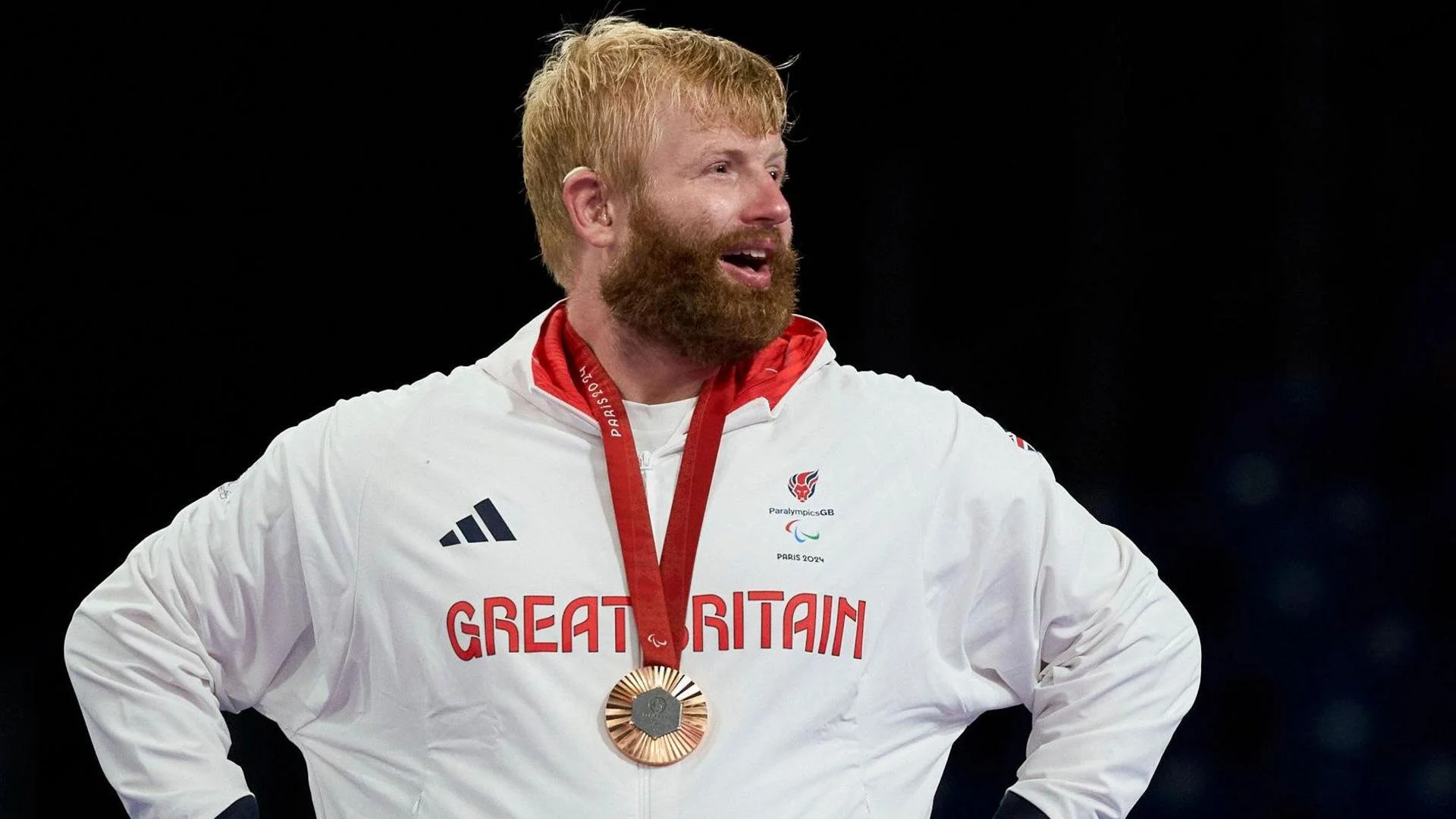 A male athlete with red hair and beard smiling, wearing a white Great Britain jacket with a bronze medal around his neck, during an award ceremony at the Paralympics.
