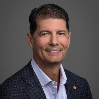 A professional headshot of a middle-aged man wearing a dark checkered blazer and light blue shirt, smiling against a gray background.