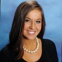 Professional portrait of a young woman with brown hair, smiling, wearing a black top and pearl necklace, against a blue background.