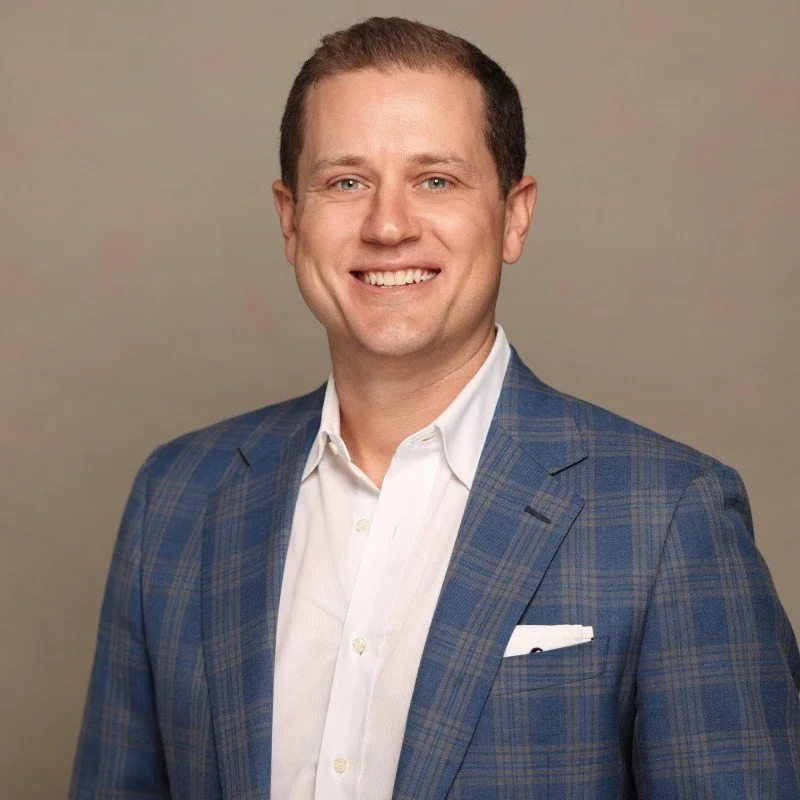 A smiling man in a blue plaid suit jacket and white dress shirt posing against a plain background.