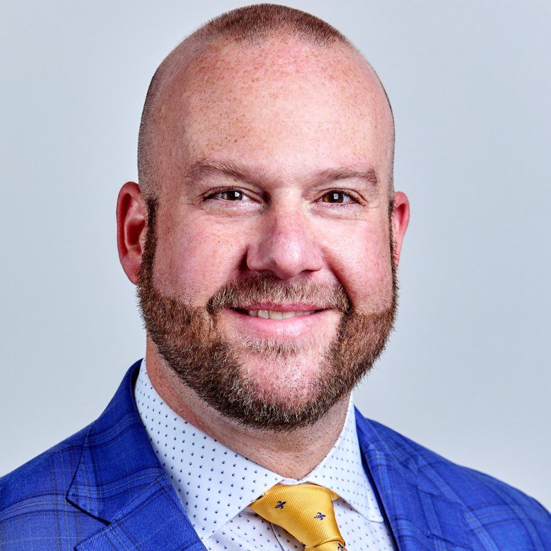 Headshot of a smiling man with a beard, wearing a blue suit, white shirt with small polka dots, and yellow tie, against a plain light gray background.