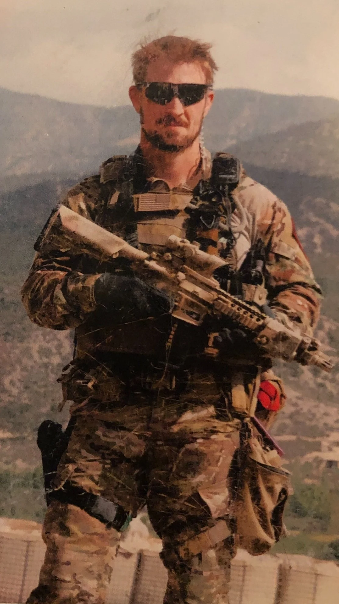 A soldier in camouflage uniform holding a rifle, wearing sunglasses, standing outdoors with mountainous terrain in the background.