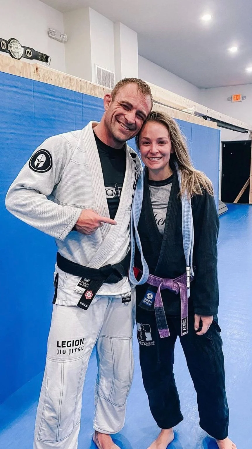 Two smiling women in Brazilian Jiu-Jitsu gis with belts, standing on a blue mat in a gym, celebrating after a match or training session.