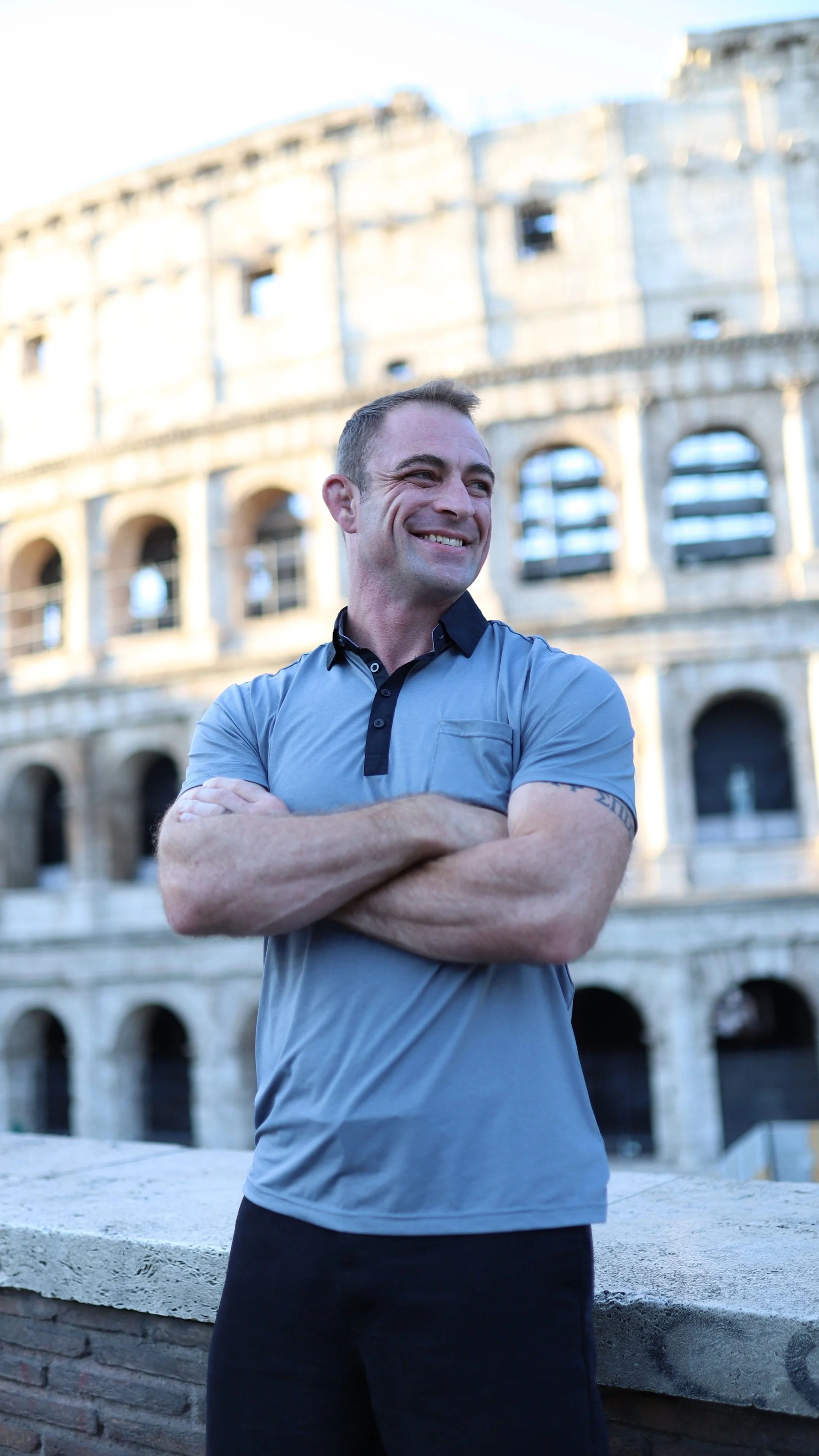 A smiling man with crossed arms standing in front of the Colosseum in Rome, Italy.