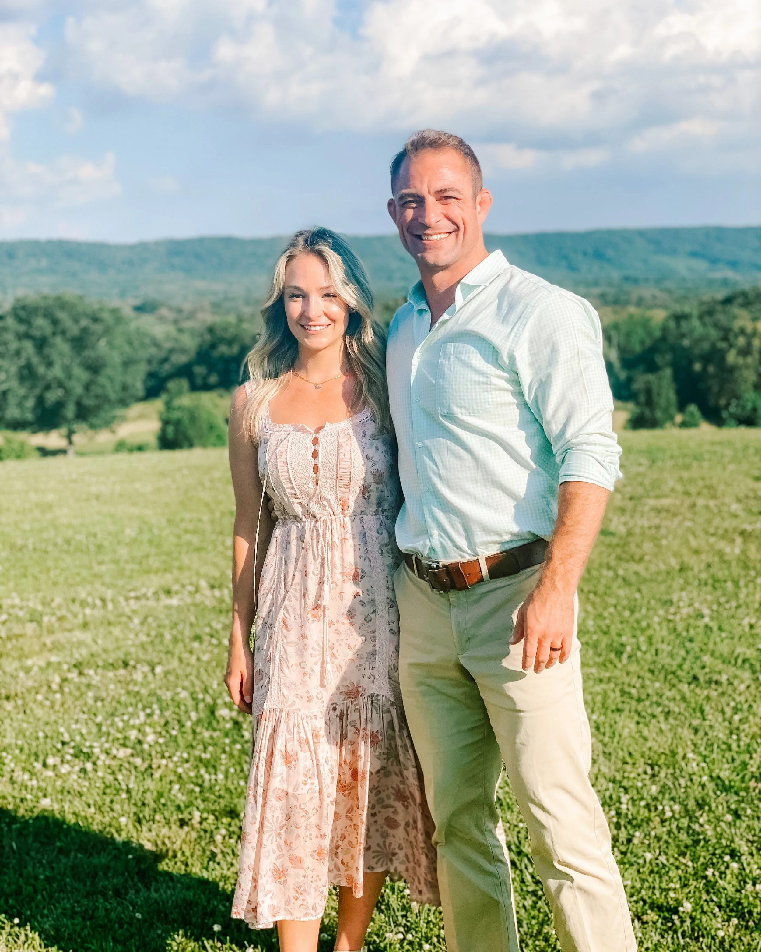 A couple standing outdoors in a grassy field with green hills and blue sky in the background, smiling at the camera.