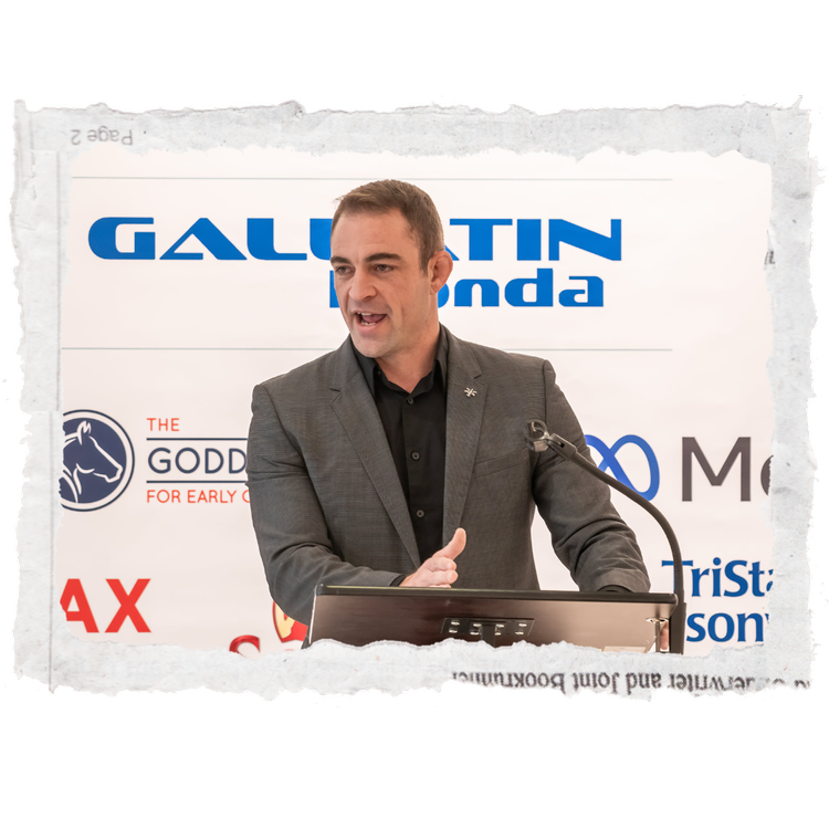 A man in a gray suit speaking at a podium with raised hand. Behind him is a banner with multiple logos including 'Gallatin,' 'Honda,' 'The Godd,' and others.