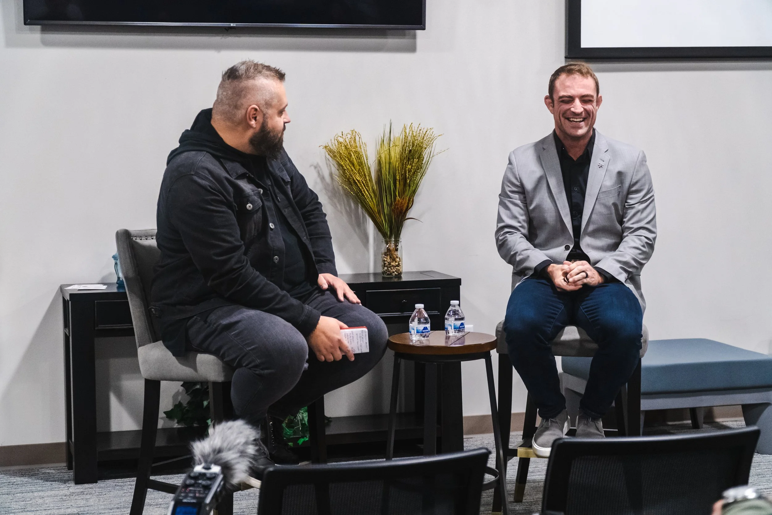 Two men sitting on chairs, engaged in a conversation. One in a black jacket, holding a piece of paper, and the other in a gray blazer, smiling. A small table with water bottles and a decorative plant are between them.
