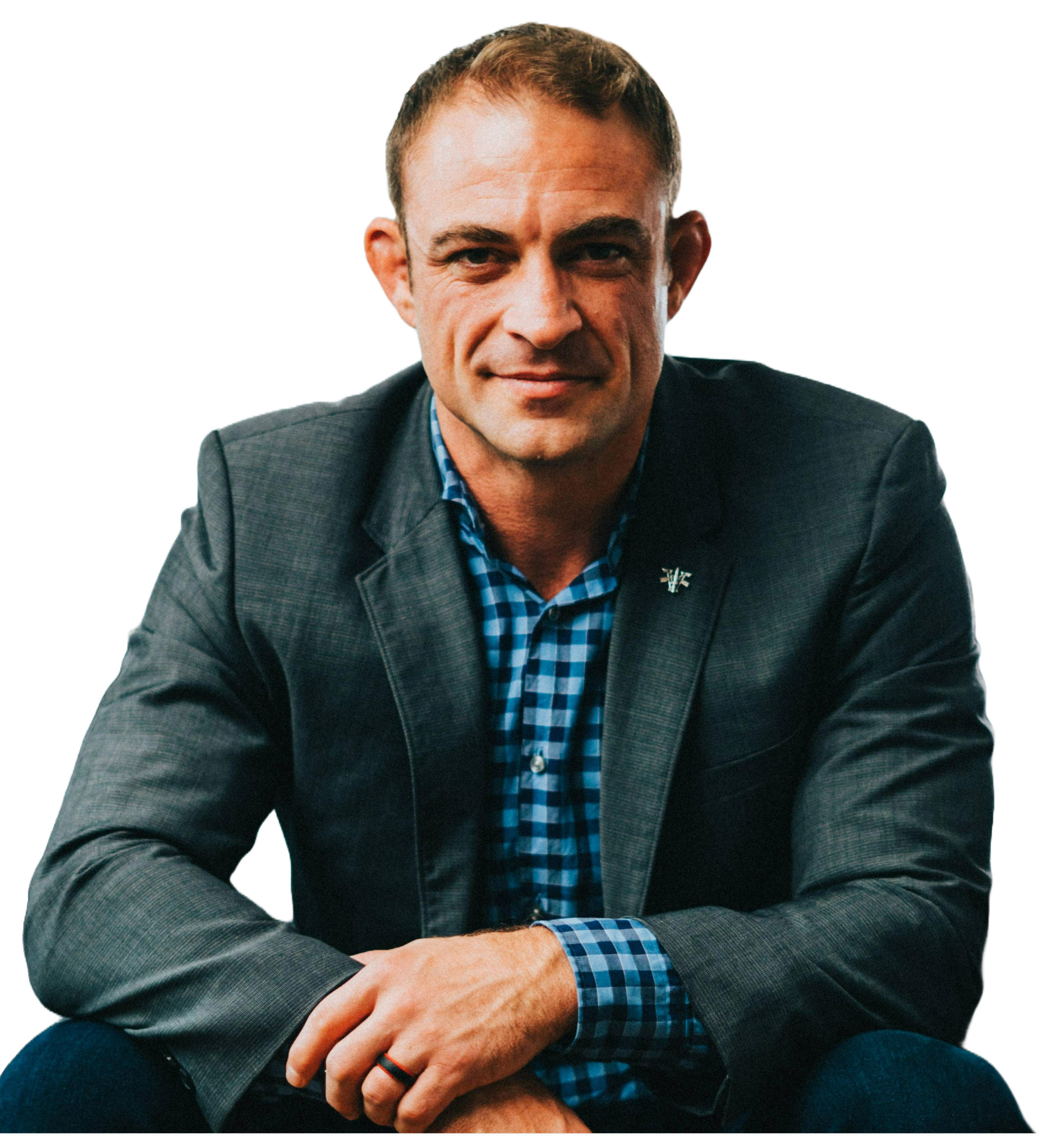 A male leadership consultant and military entrepreneur wearing a dark gray blazer and blue checkered shirt, sitting with his hands crossed, smiling slightly.