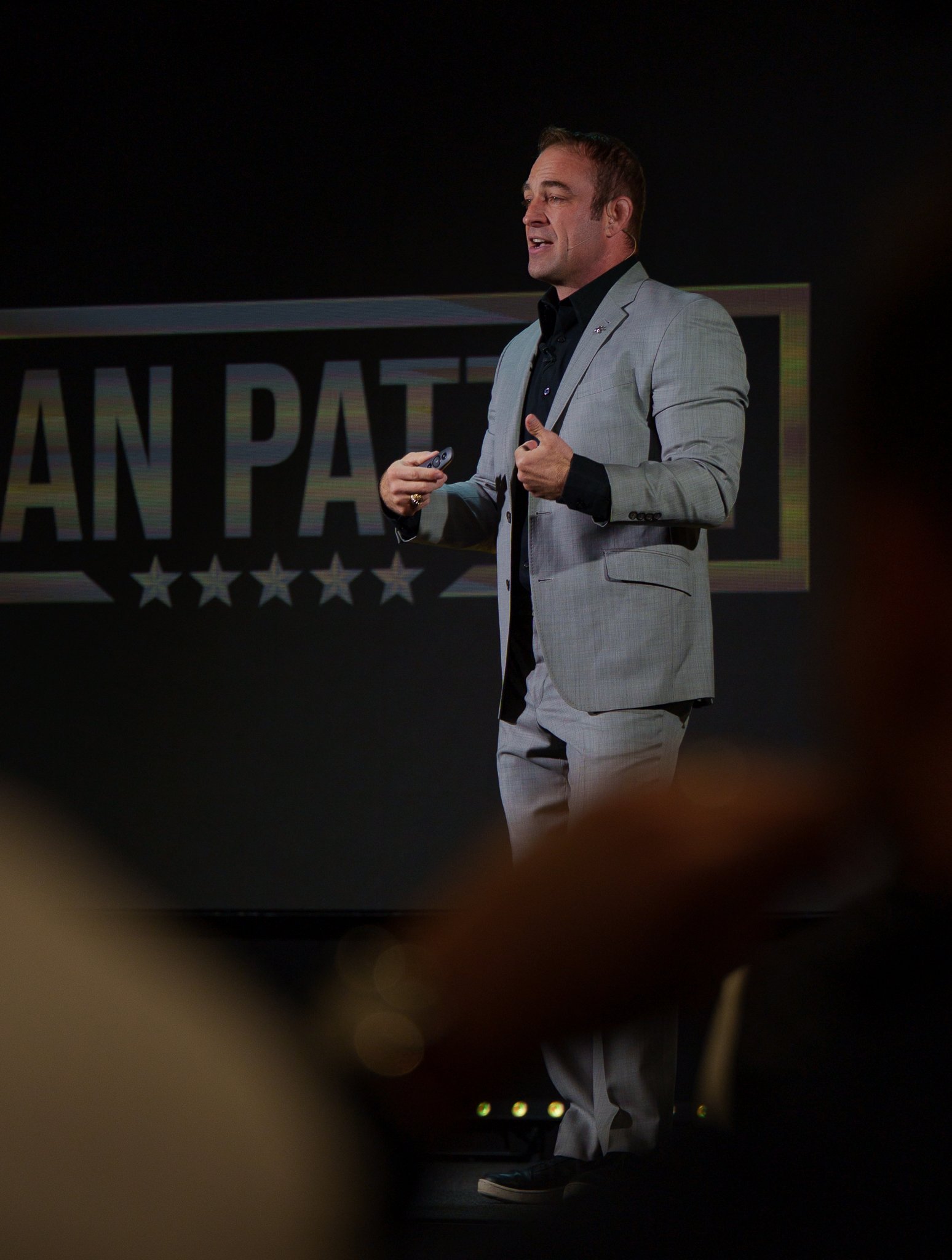 A man in a light gray suit giving a presentation on stage, standing in front of a screen with the text "AN PATR" and stars beneath it.