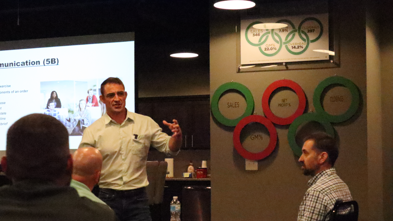A man in a light shirt giving a presentation to a group of people in a conference room. There are charts and diagram boards on the wall behind him, with one showing sales data, gross profit, net profit, and overhead percentages. Some attendees are visible, seated and listening.