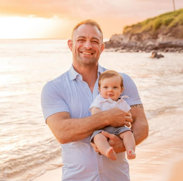 A man smiling and holding a baby on the beach with a sunset and shoreline in the background.
