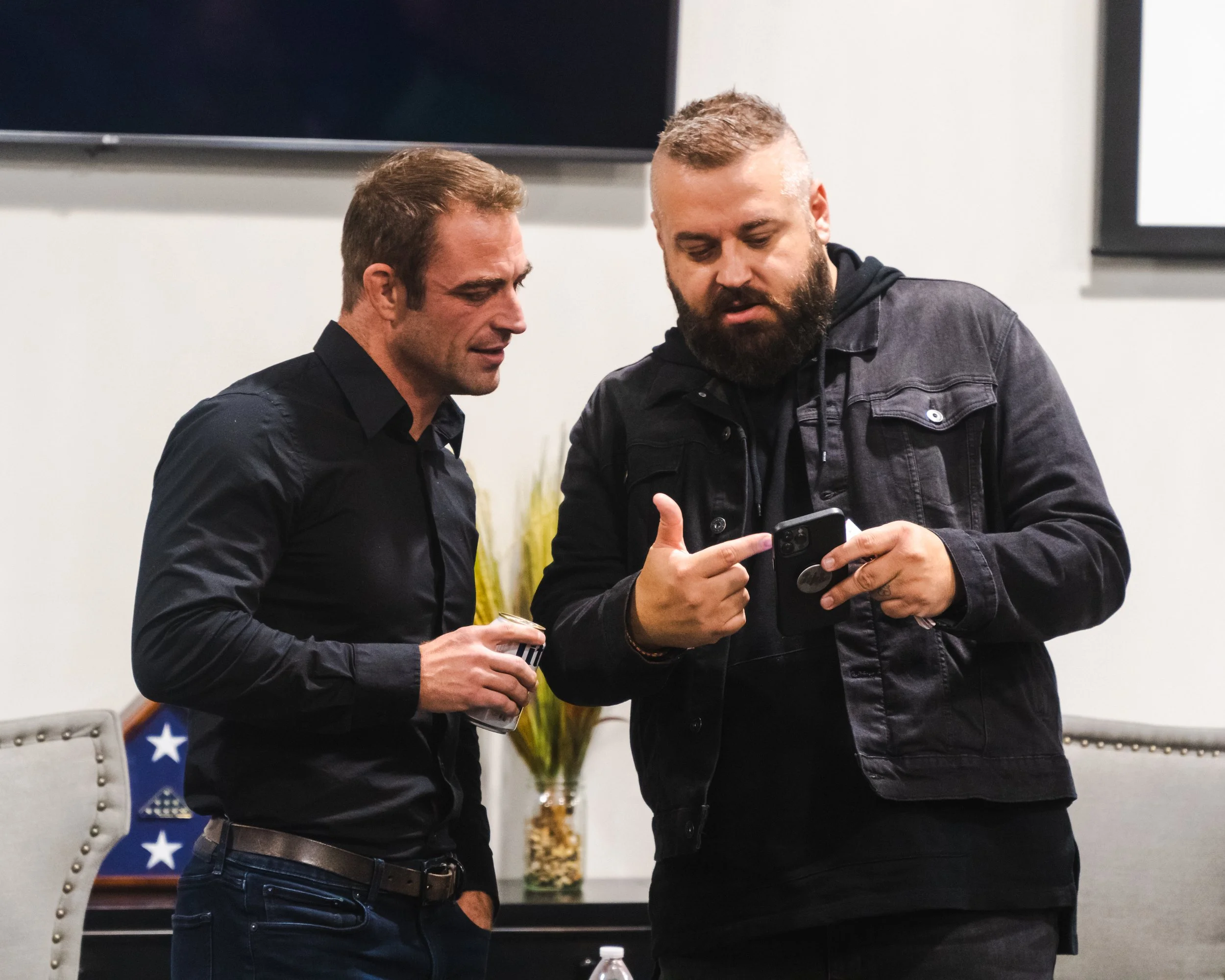 Two men standing close together, looking at a smartphone. One man is holding a drink, and the other is pointing at the phone screen.