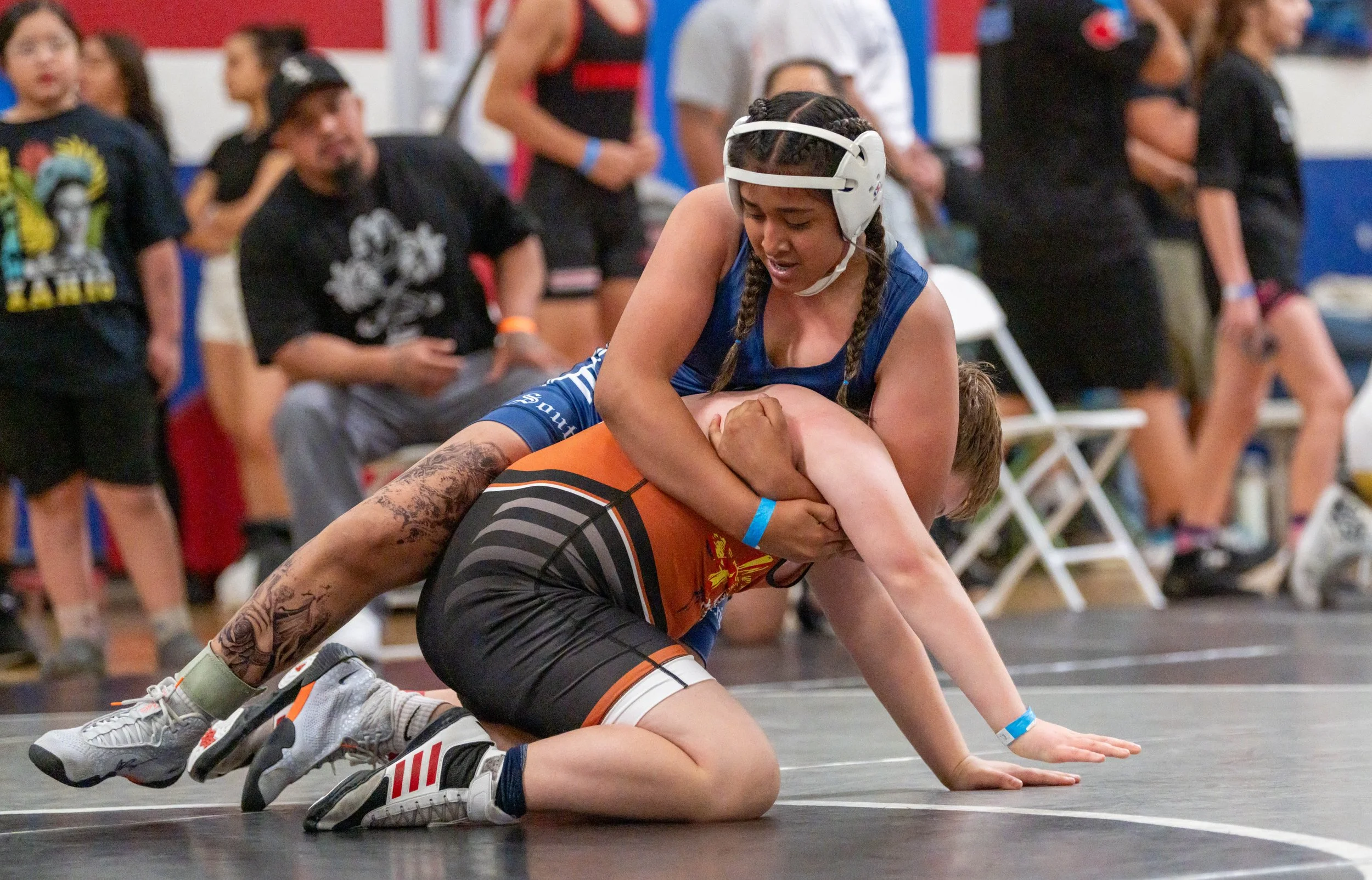 Two female wrestlers competing on a wrestling mat; one wearing blue and the other in orange and black, with the wrestler in blue on top, pinning her opponent.