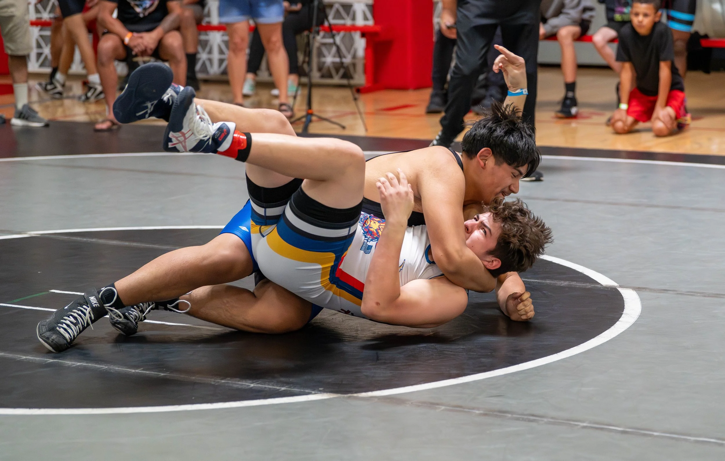 Two young male wrestlers competing in a match on a black wrestling mat. One wrestler is on top, pinning the other to the mat, with spectators watching in the background.